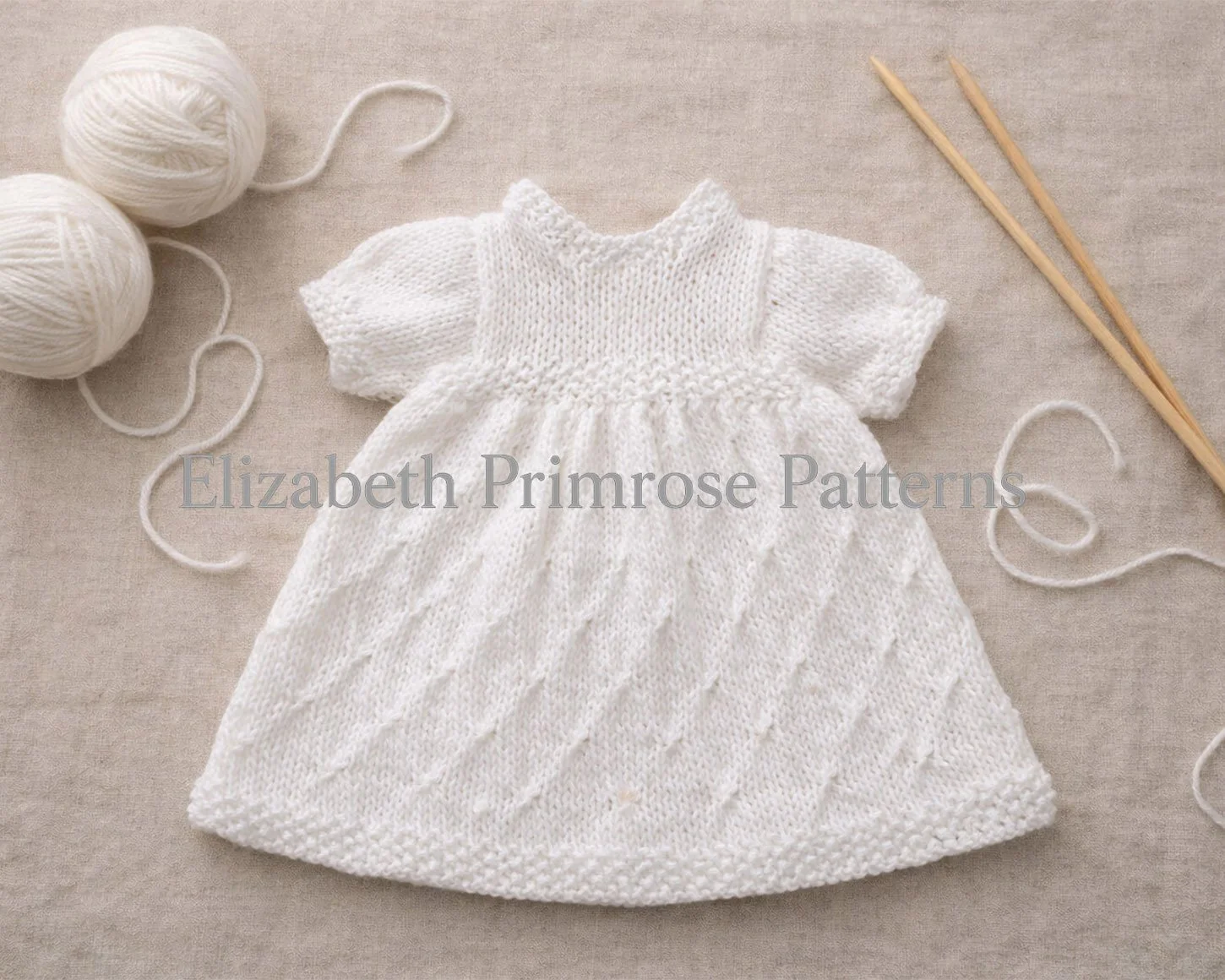 White hand knitted doll dress laid flat on beige linen with textured bodice, diamond skirt pattern, yarn balls and wooden needles beside it.