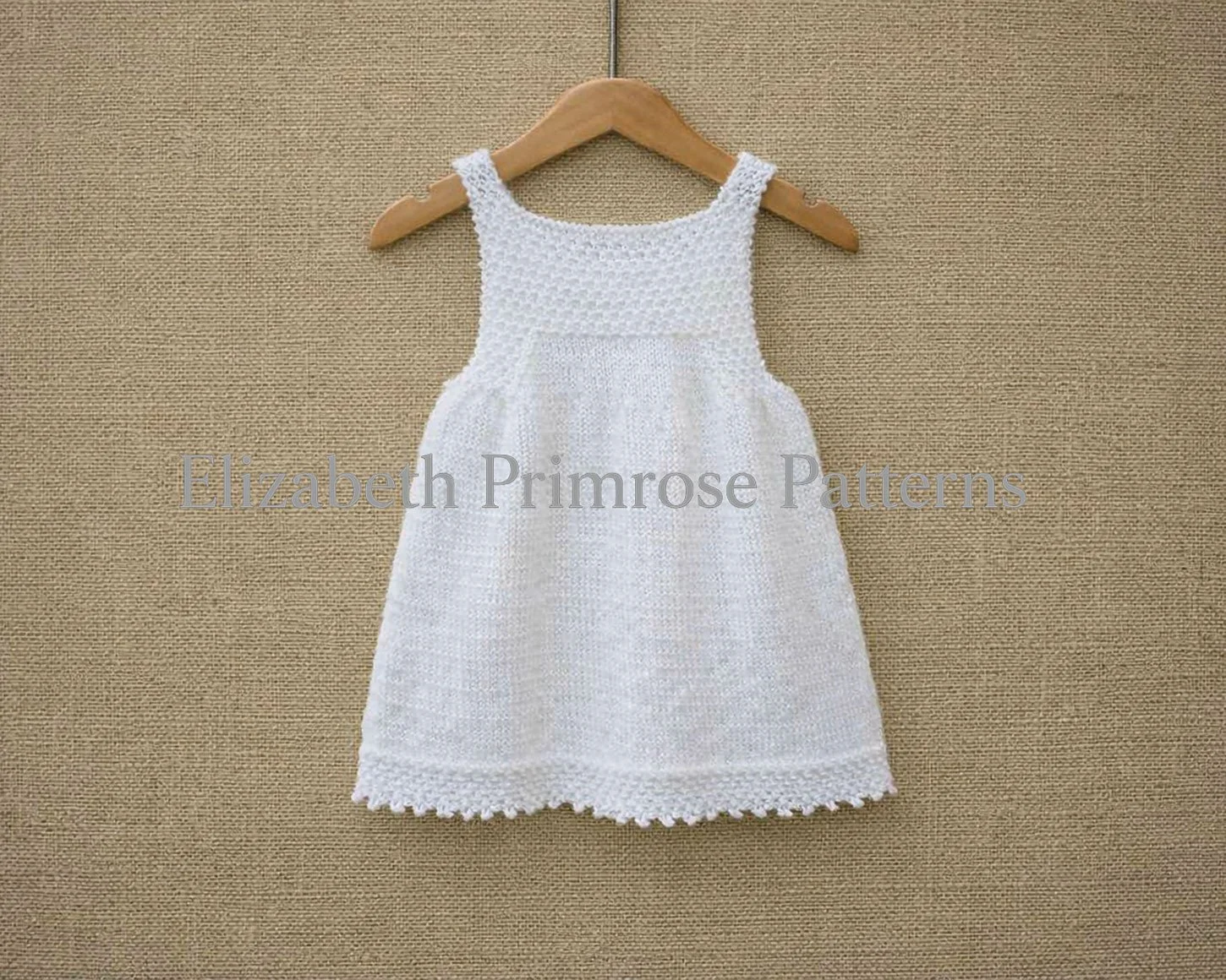 Back view of white hand knitted children’s pinafore with textured bodice, straps and smooth skirt, hanging on a wooden hanger against a fabric backdrop.