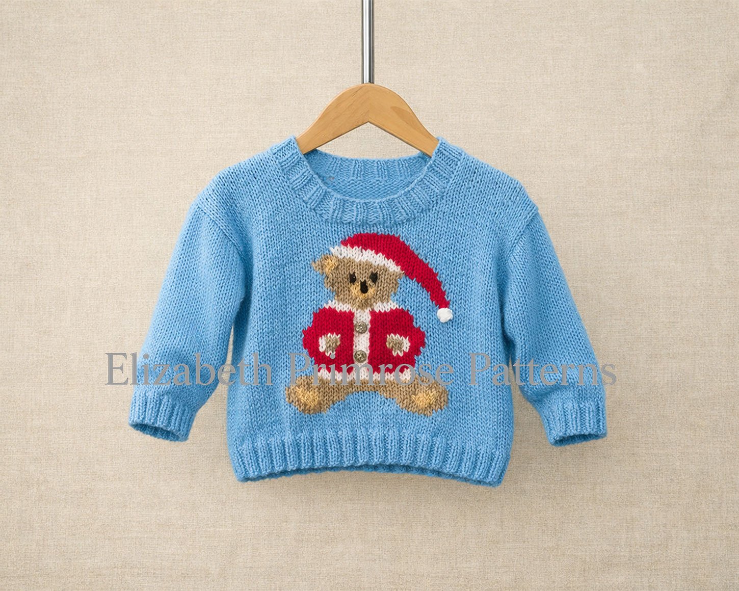 Hand knitted blue child’s sweater with a Santa clad teddy bear motif, shown on a wooden hanger against a neutral beige fabric backdrop.