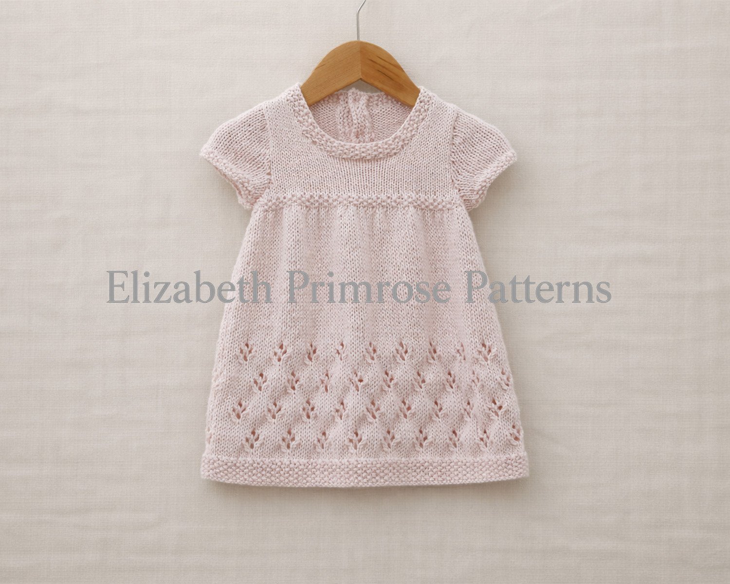 Light pink hand knitted baby dress with short sleeves, hanging on a wooden hanger against an uncreased light beige linen flatlay background.
