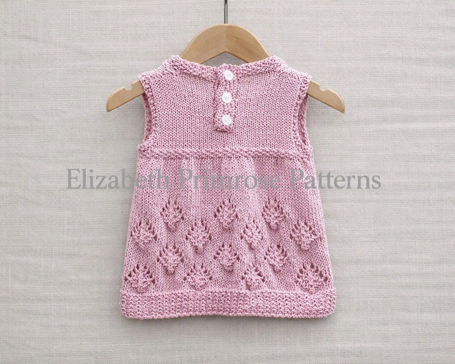 Sleeveless pink knitted baby dress with pinecone pattern skirt hanging from a wooden hanger against a light beige linen backdrop, seen from the back view.
