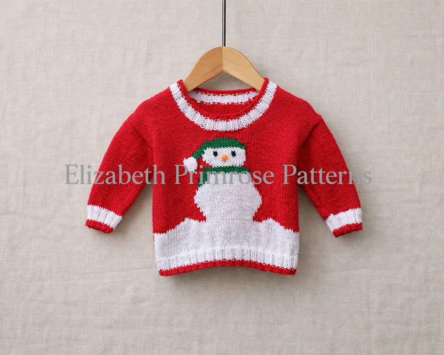 Red hand knitted children’s jumper with white cuffs and collar, featuring a snowman motif wearing a green hat, hanging on a wooden hanger from a vertical metal rod against a linen background.
