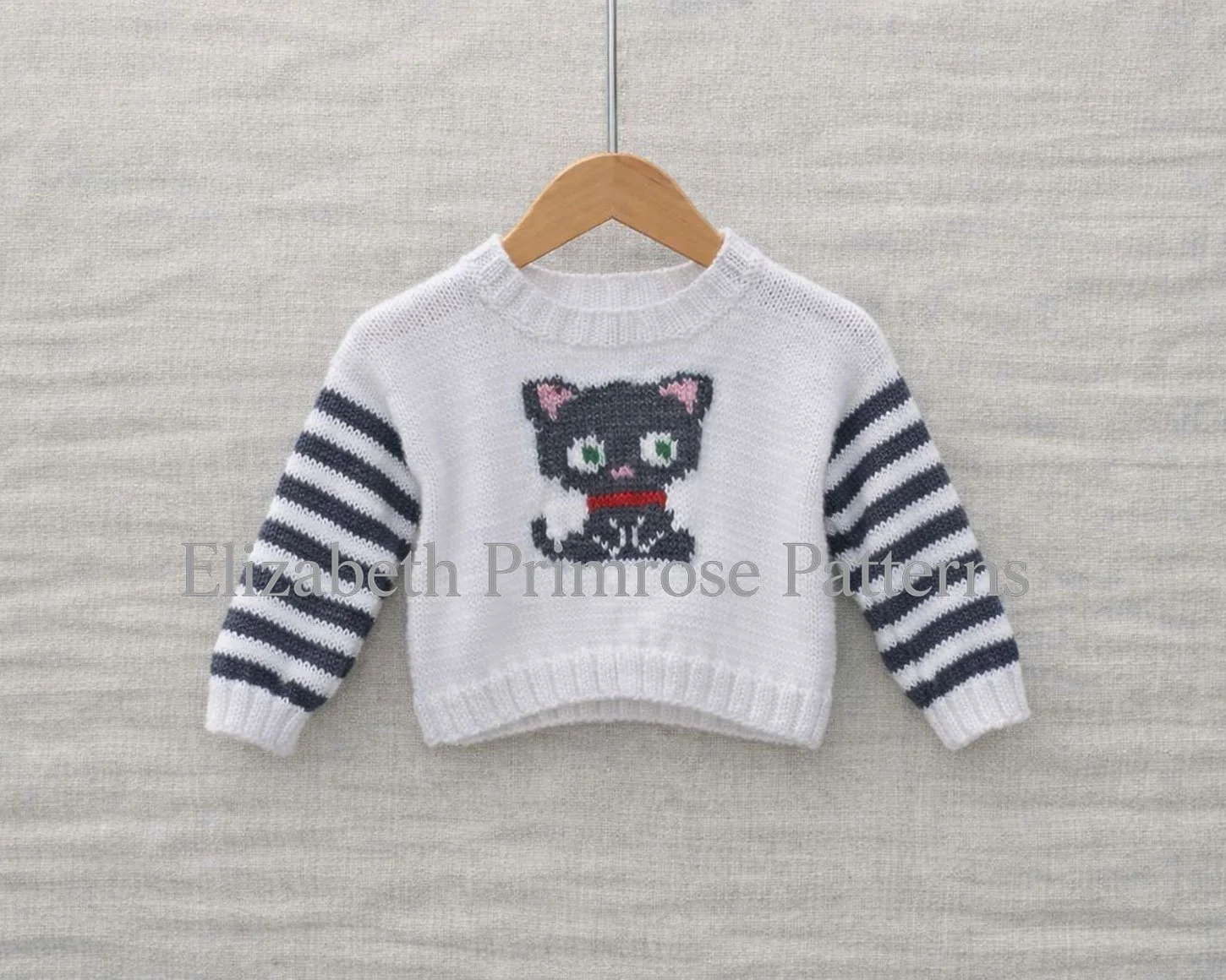 White baby jumper with striped sleeves and a knitted grey cat motif hanging on a wooden hanger against a soft, neutral background.