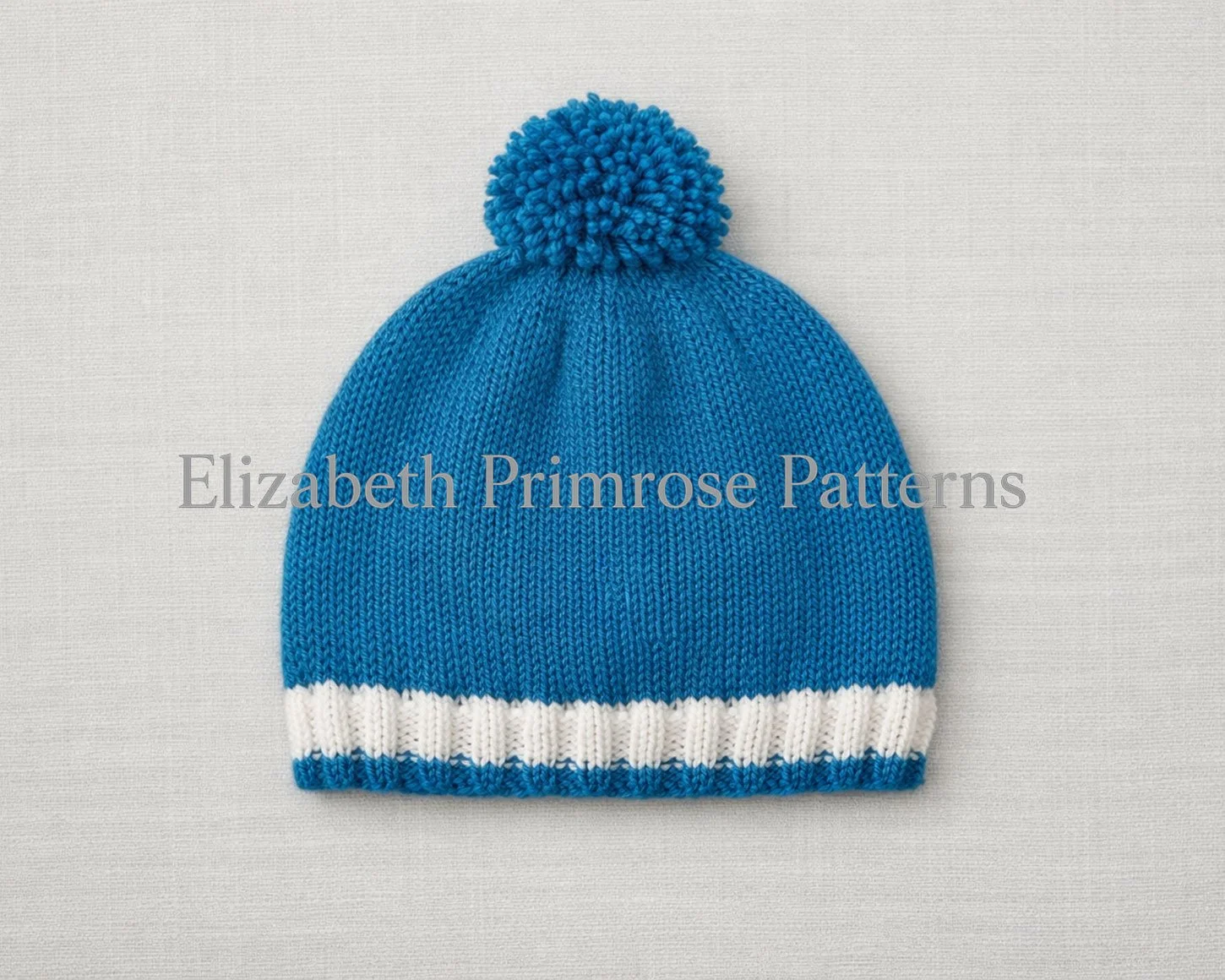 Knitted blue children’s hat with pompom, laid flat on a soft neutral background