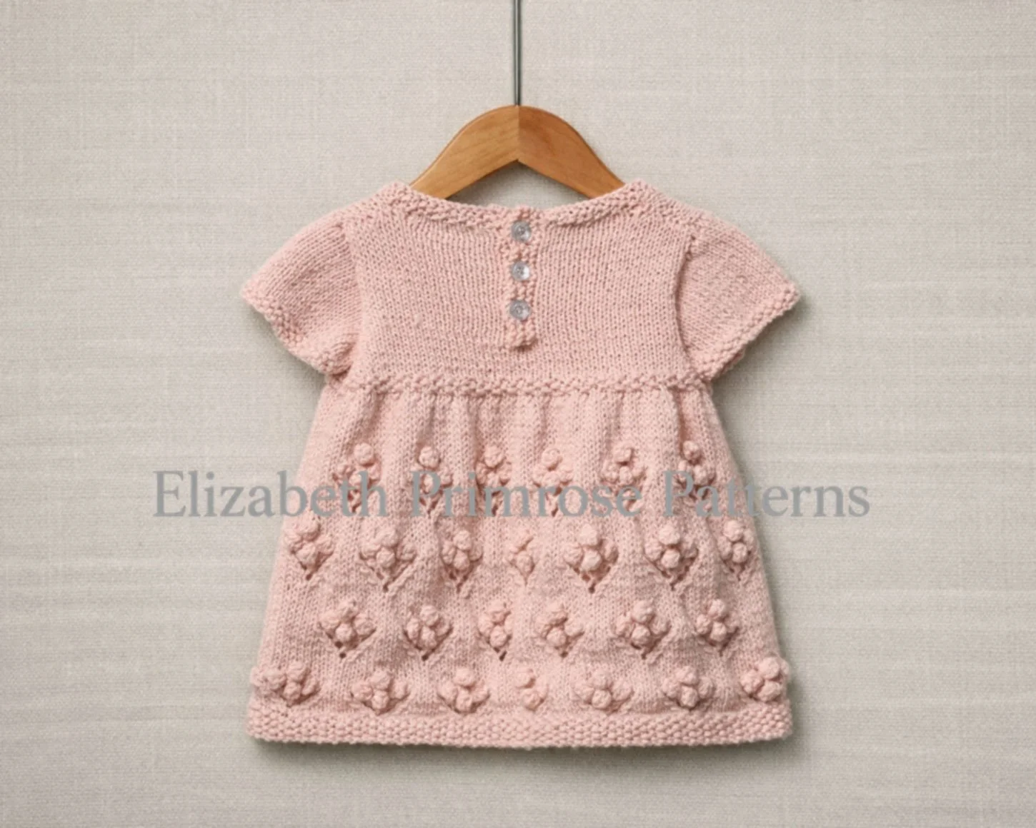 Back view of pink hand knitted short-sleeved children’s dress with bobble flower skirt, 3 button fastening, hanging on a wooden hanger and shown against a neutral fabric background.