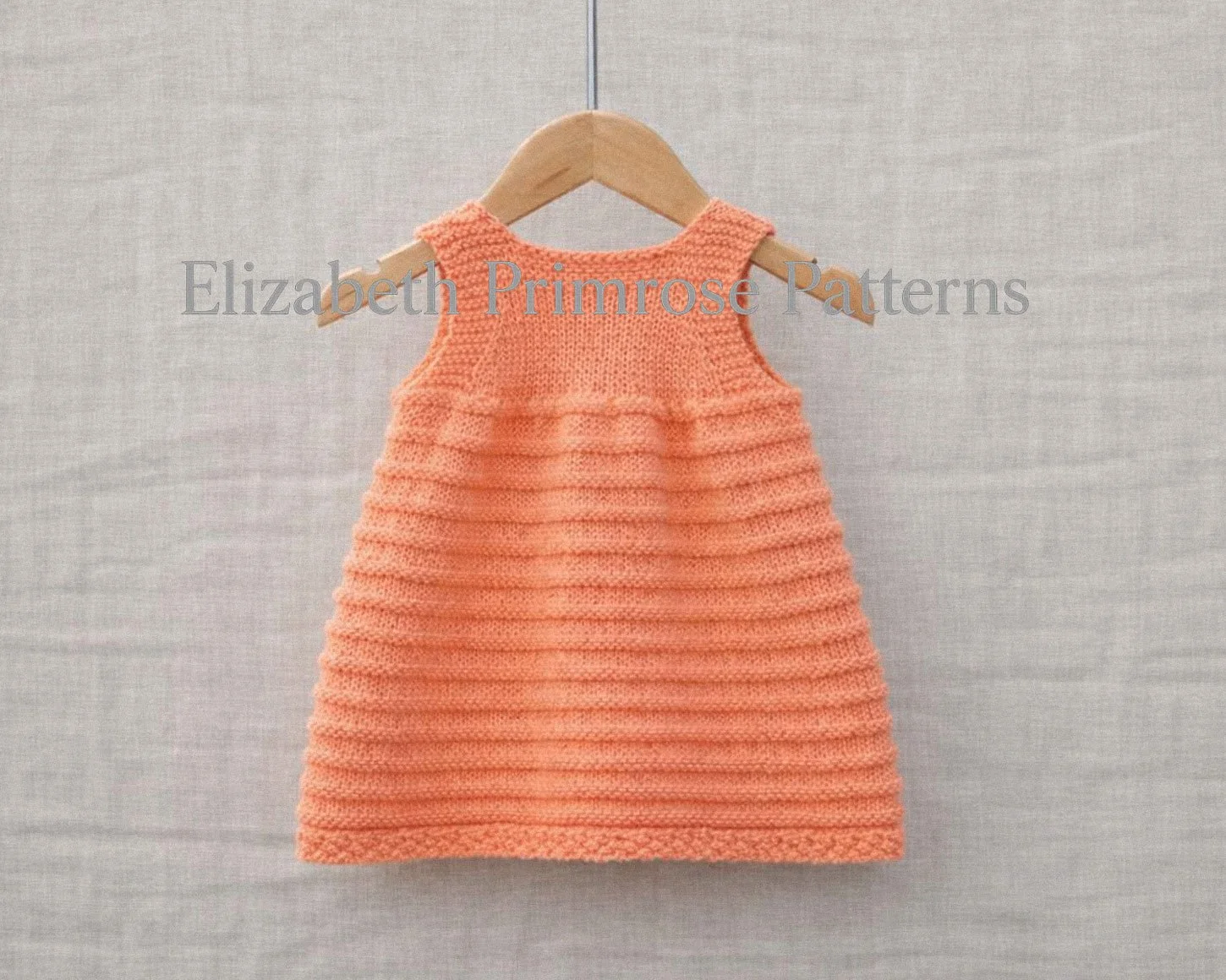 Back view of hand‑knitted peach baby pinafore with textured ridges and bobbles displayed on a hanger over a neutral fabric backdrop.