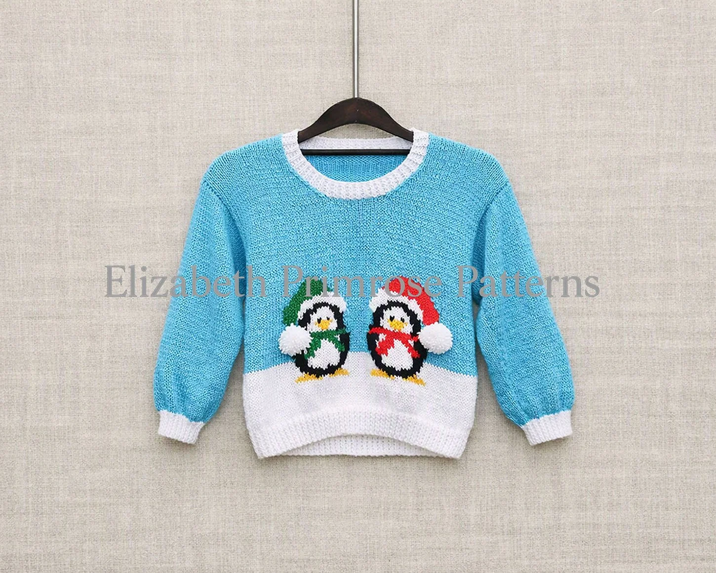 Light blue children’s jumper with two penguins in winter hats, hanging on a wooden hanger against a smooth beige linen background.