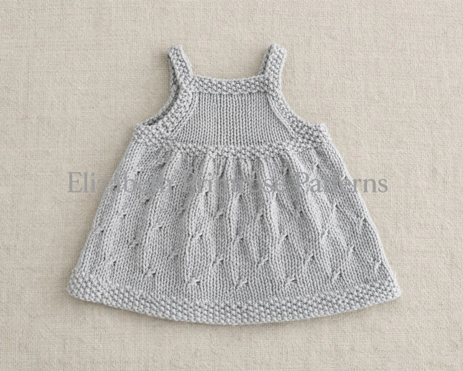Back view of beige dolls dress displayed on linen backdrop, featuring diamond textured skirt and moss stitch borders.