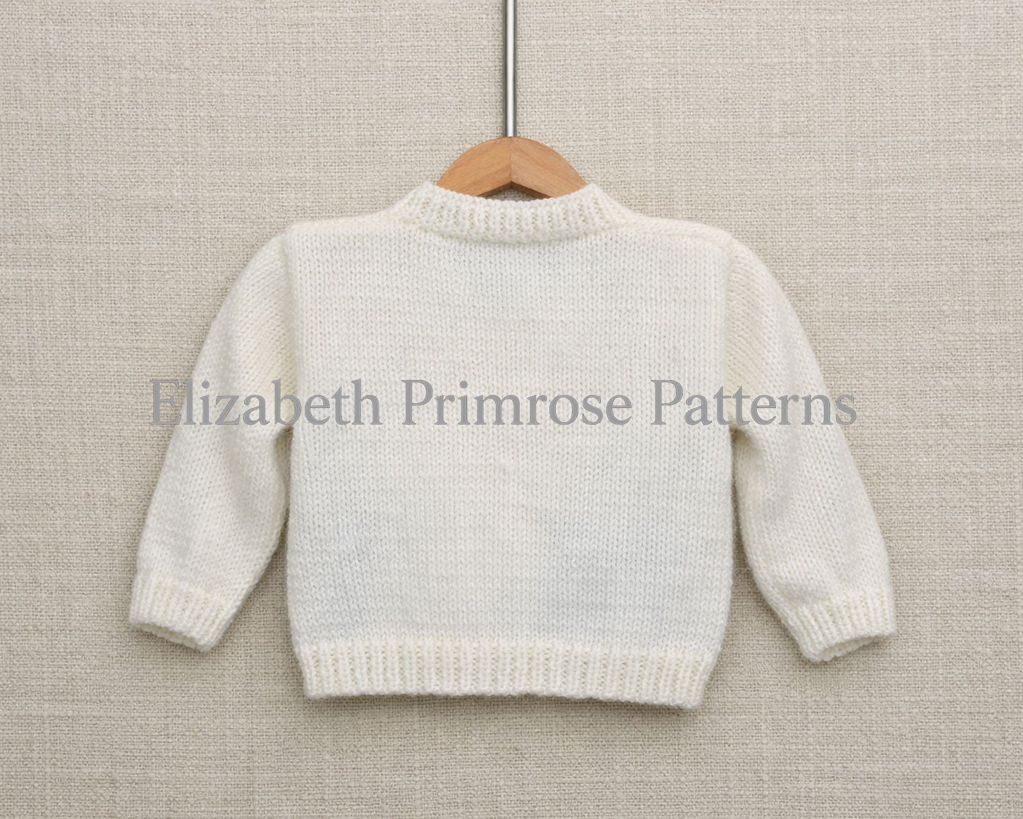 Cream knitted baby cardigan shown from the back on a wooden hanger against a beige linen background, highlighting the simple ribbed neckline and cuffs.