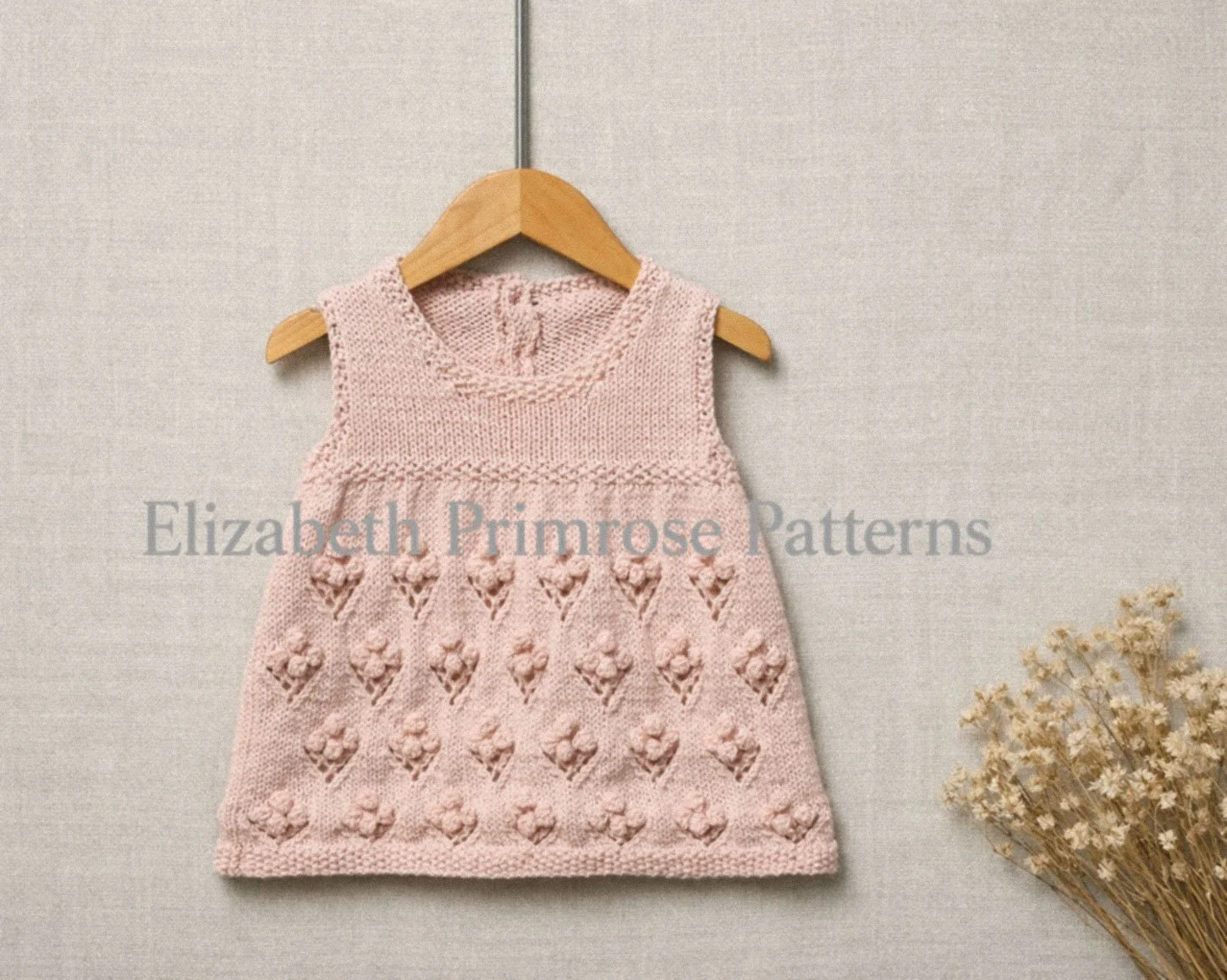 Pink hand knitted sleeveless baby dress with bobble flower skirt hanging on a wooden hanger, shown against a soft neutral fabric background with dried flowers.