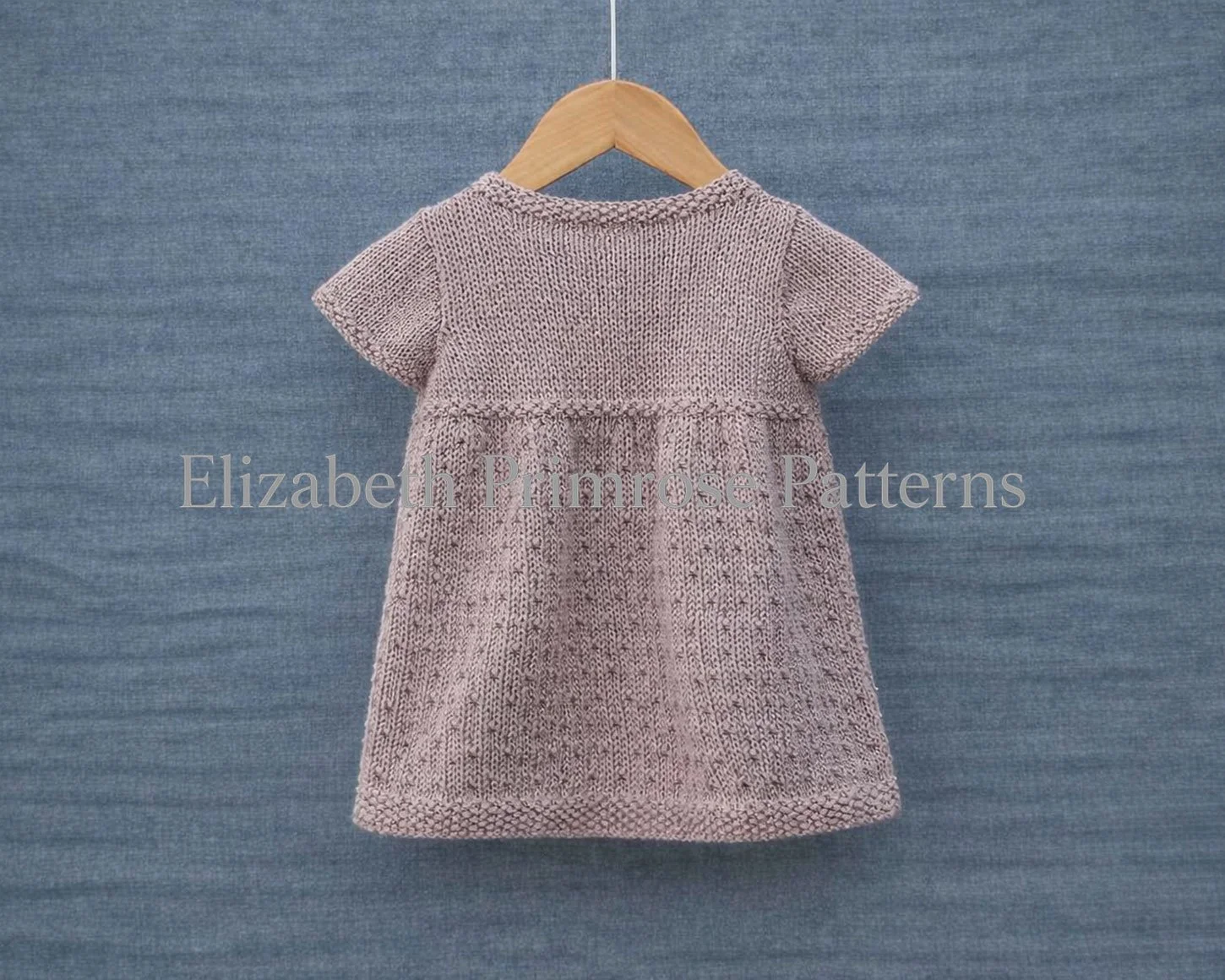 Back view of short sleeved mauve dress featuring a textured skirt and moss stitch edgings on a hanger and blue background.