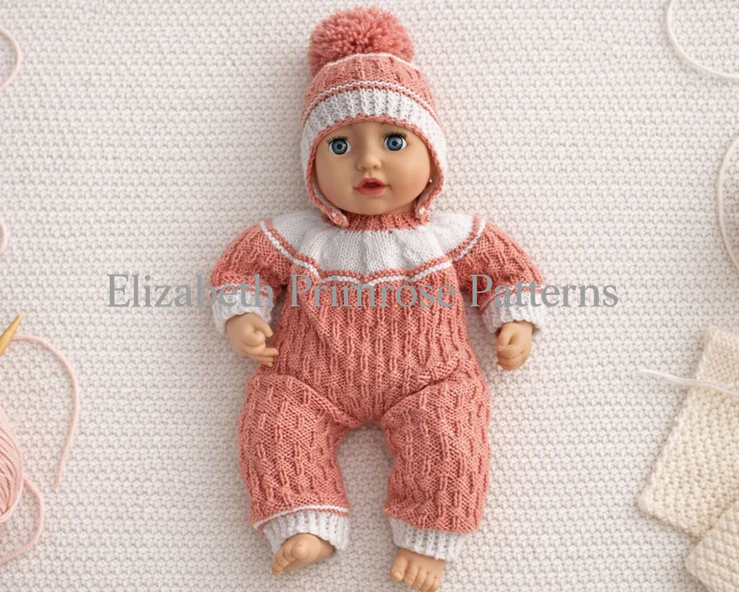 Baby doll in pink and white knitted romper and pompom hat, lying on textured cream knitted background surrounded by yarn balls and knitting needles.