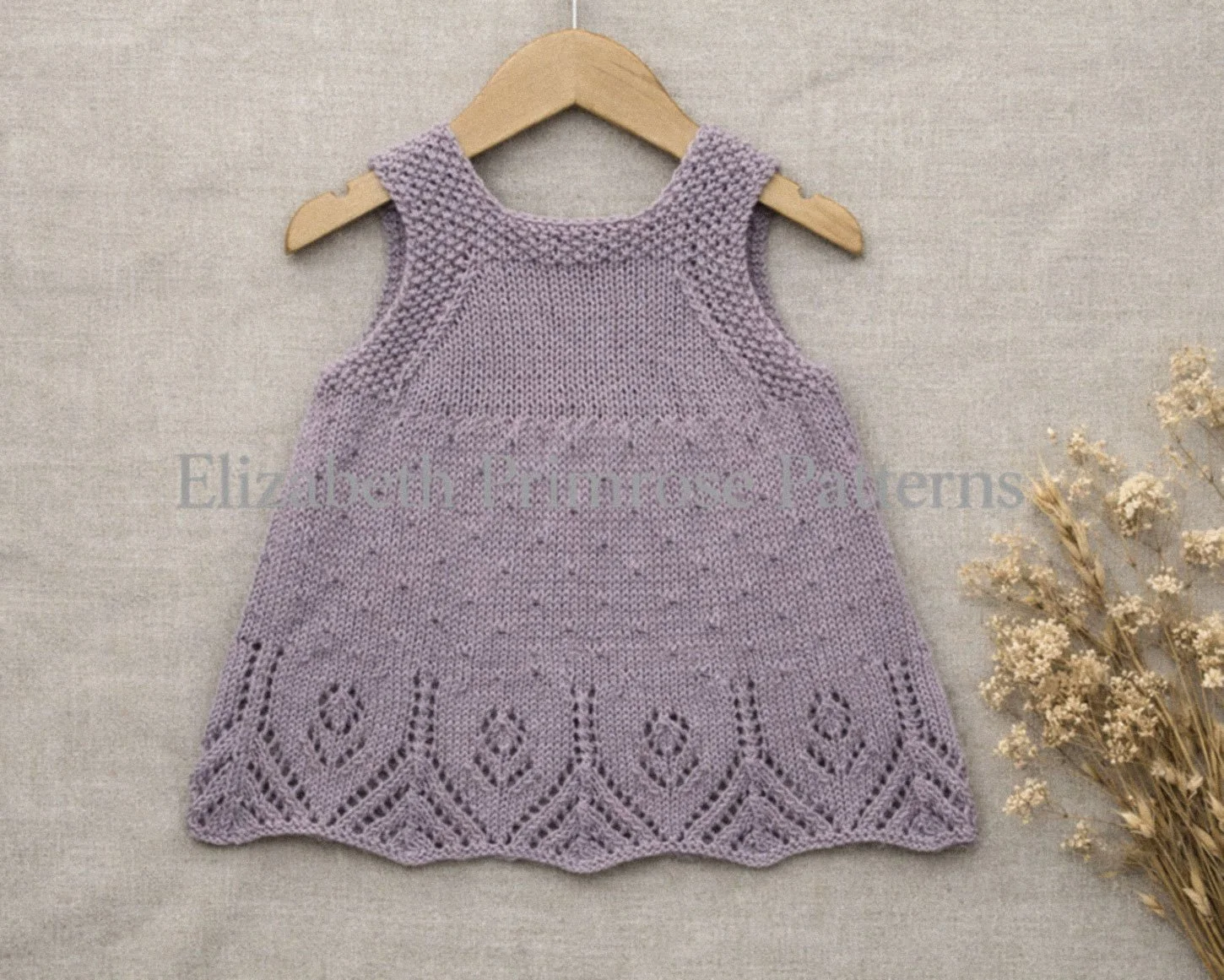 Back view of hand knitted lavender pinafore dress displayed on a hanger, showing delicate texture, lace hem and buttoned straps, set against a neutral background with dried flowers.