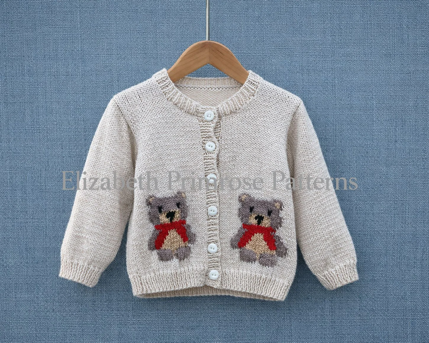 Cream hand‑knitted children’s cardigan with two beige teddy bear motifs wearing red scarves, displayed on a wooden hanger against a blue linen background.