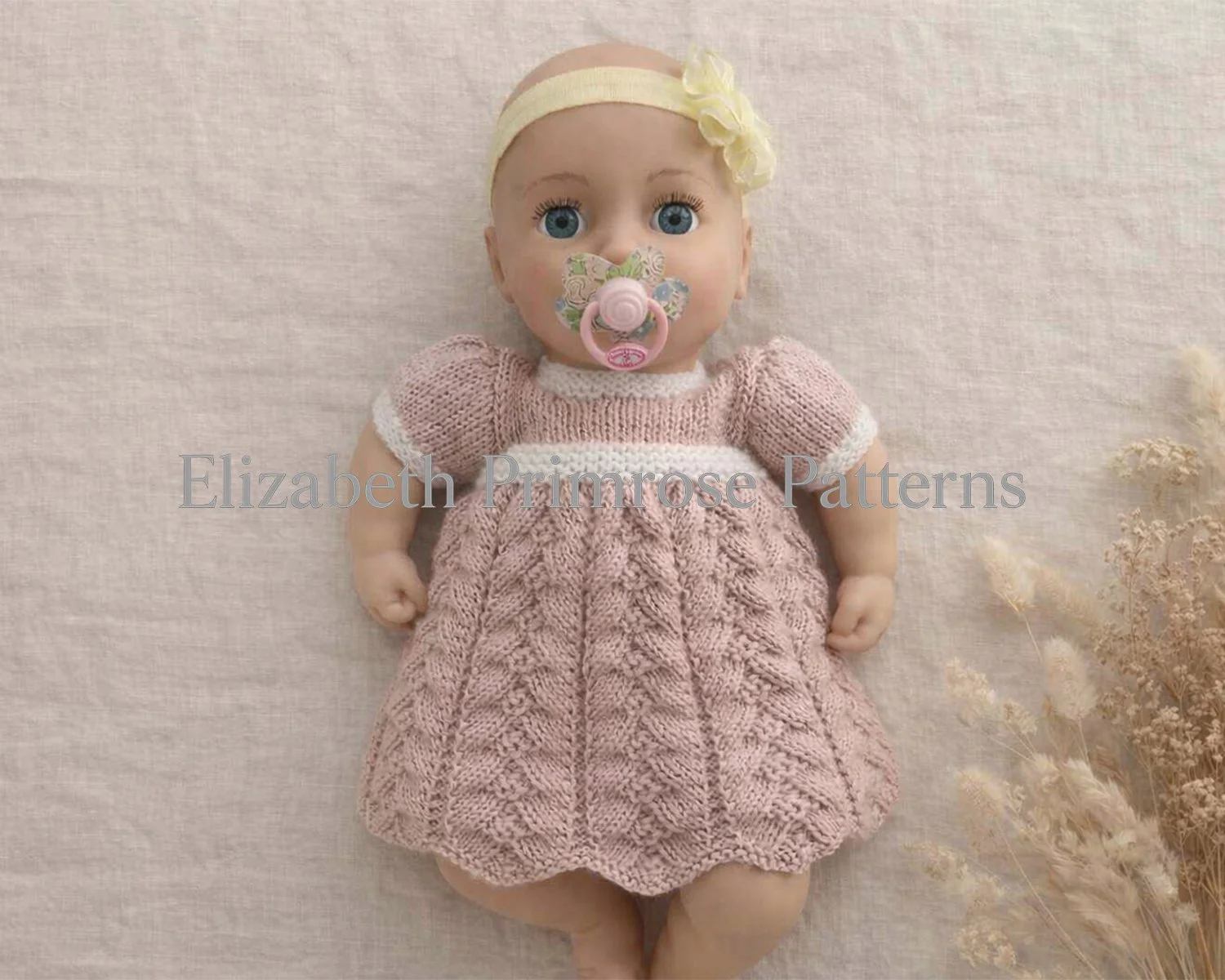 Pink herringbone doll dress on doll with pacifier on linen background, knitting pattern.
