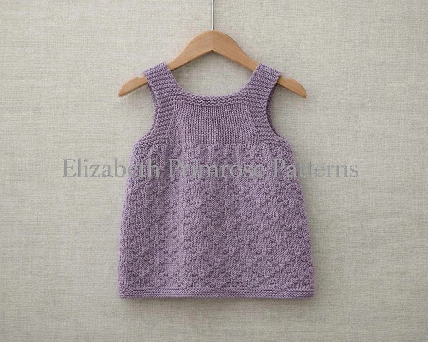Back view of textured lavender children’s pinafore dress with diamond pattern and straps displayed on a beige linen flatlay.