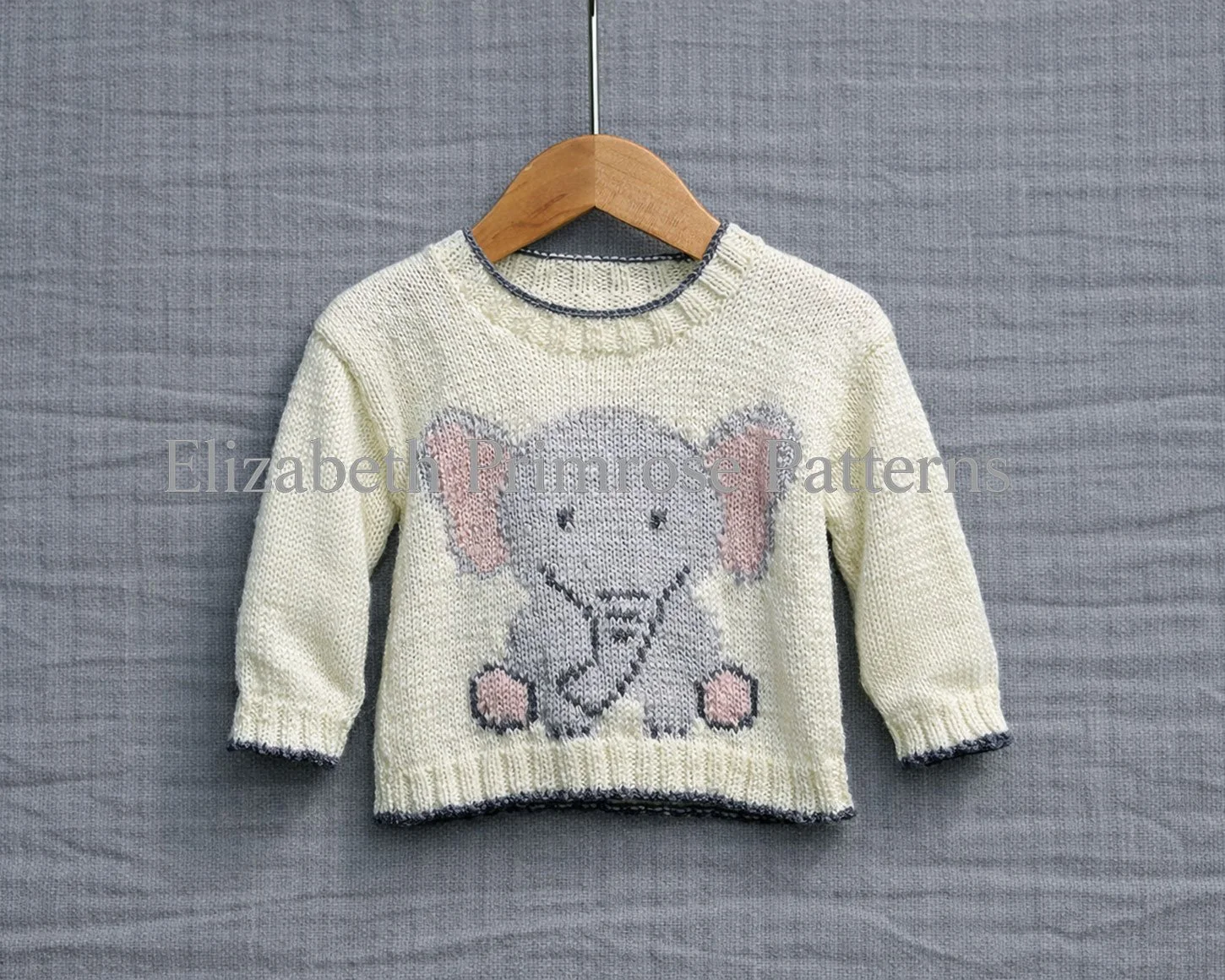 Cream knitted children’s jumper on a wooden hanger with a grey elephant motif and pink accents, shown against a sharp grey linen background.