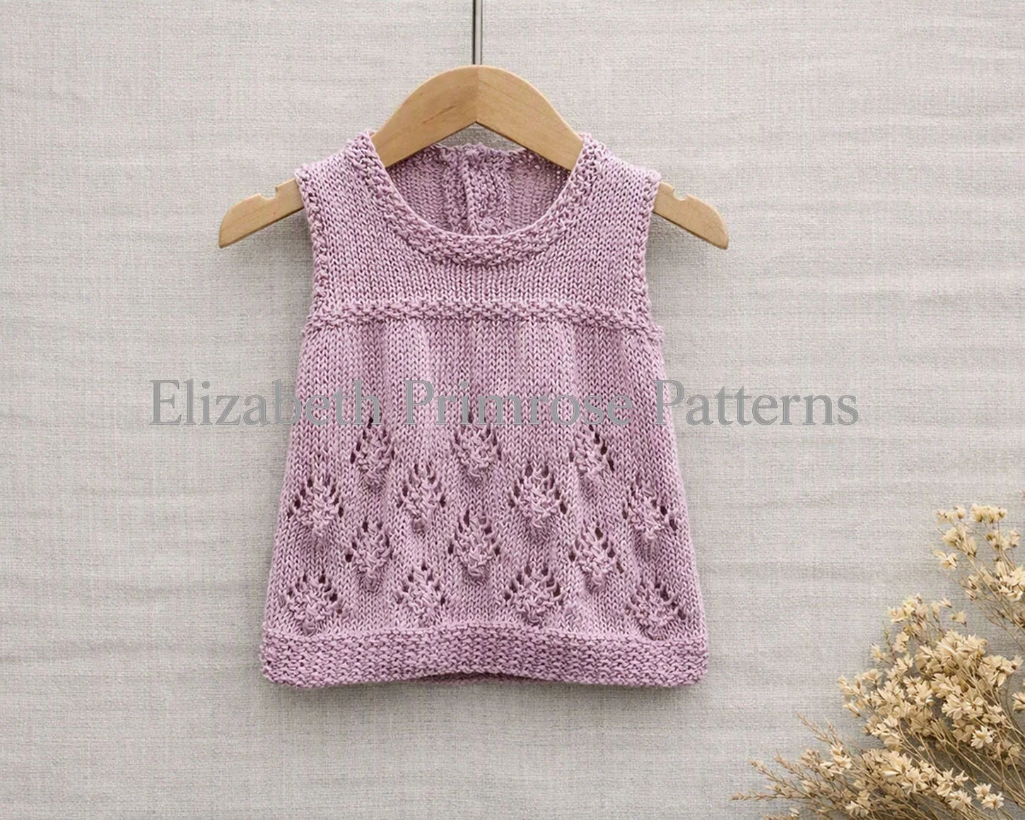 Sleeveless pink knitted baby dress with pinecone pattern skirt hanging from a wooden hanger against a light beige linen backdrop.