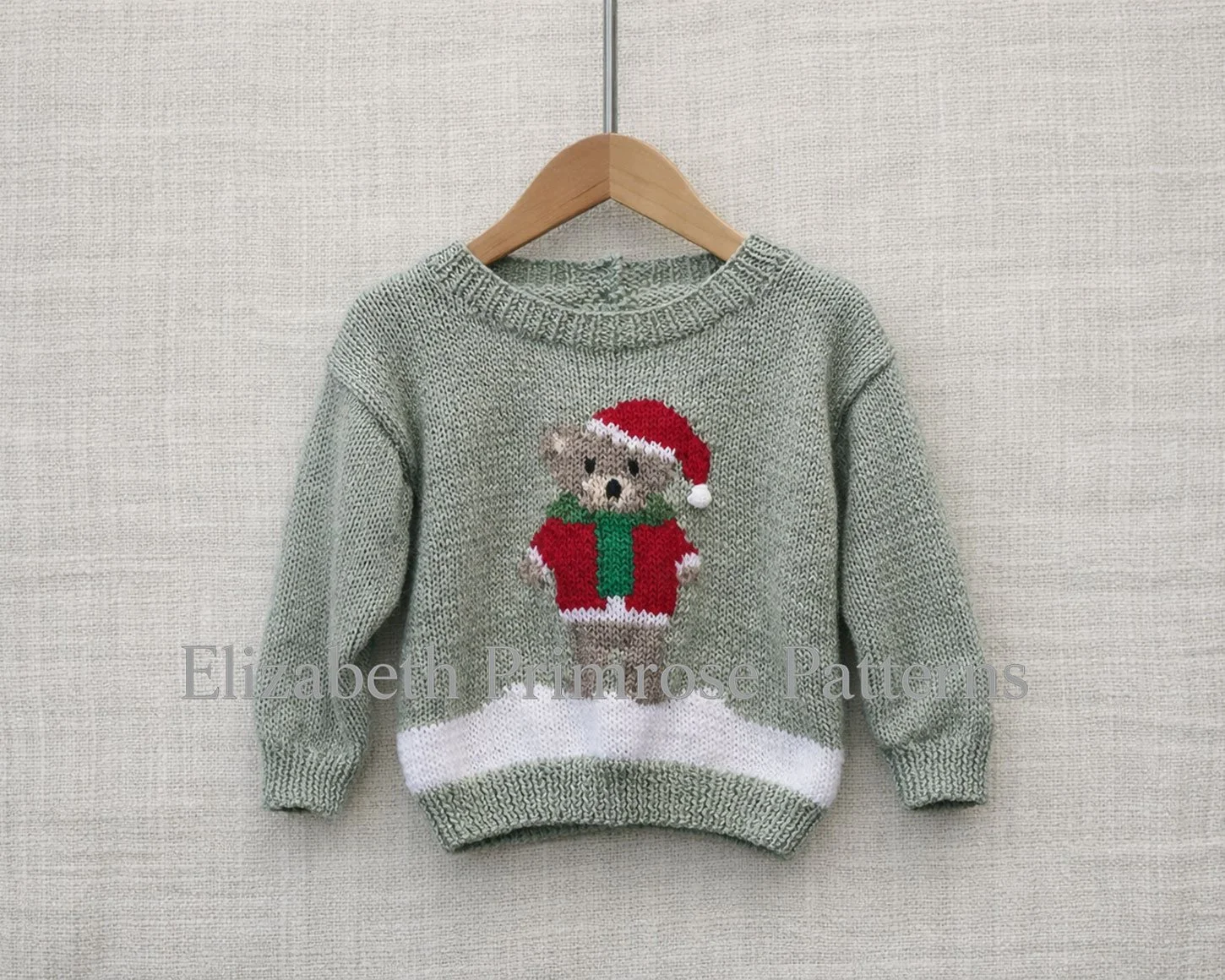 Hand‑knitted grey‑green children’s jumper featuring a teddy bear in a red Santa outfit and green scarf, hanging on a wooden hanger against a light linen backdrop.