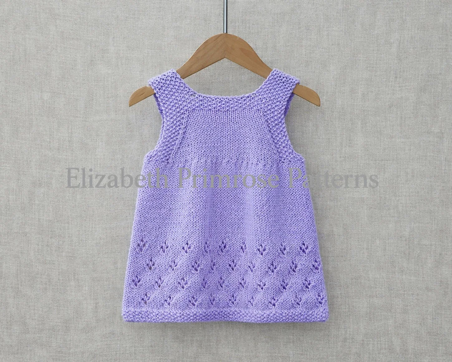 Back view of lilac hand knitted pinafore dress on a hanger with moss stitch edges and a lacy snowflake skirt against a soft fabric backdrop.