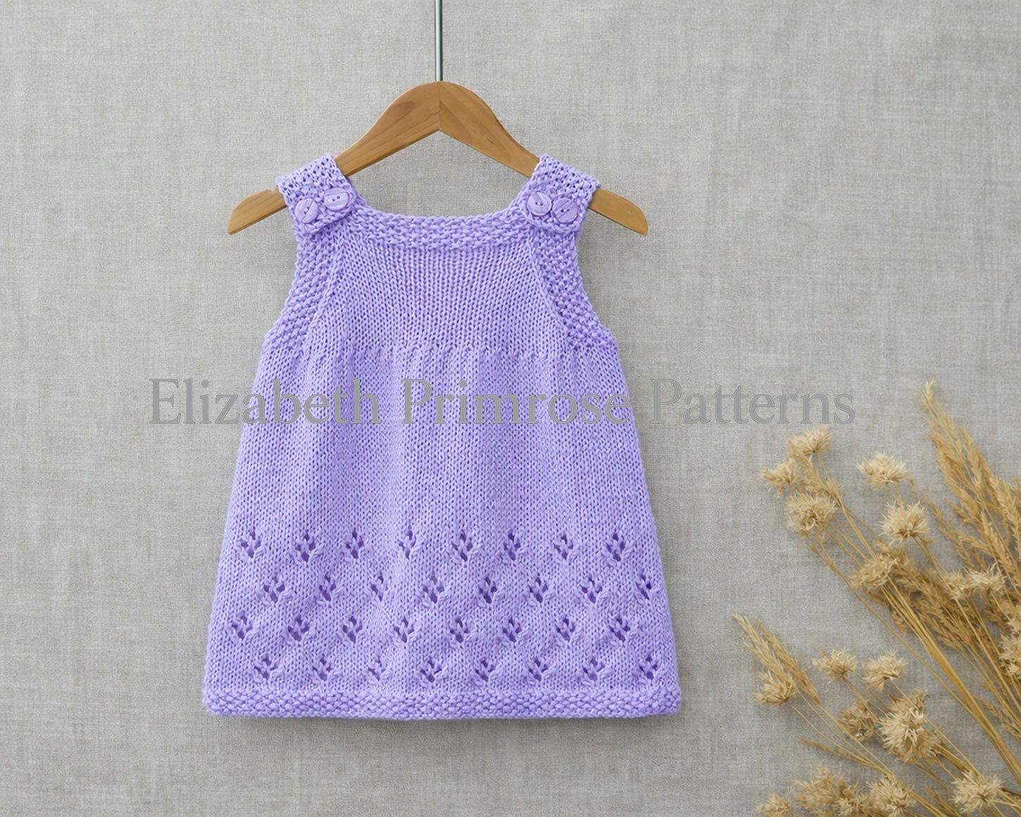 Lavender hand knitted pinafore dress on a hanger with buttoned straps, moss stitch edges, and a lacy snowflake skirt against a soft fabric backdrop.