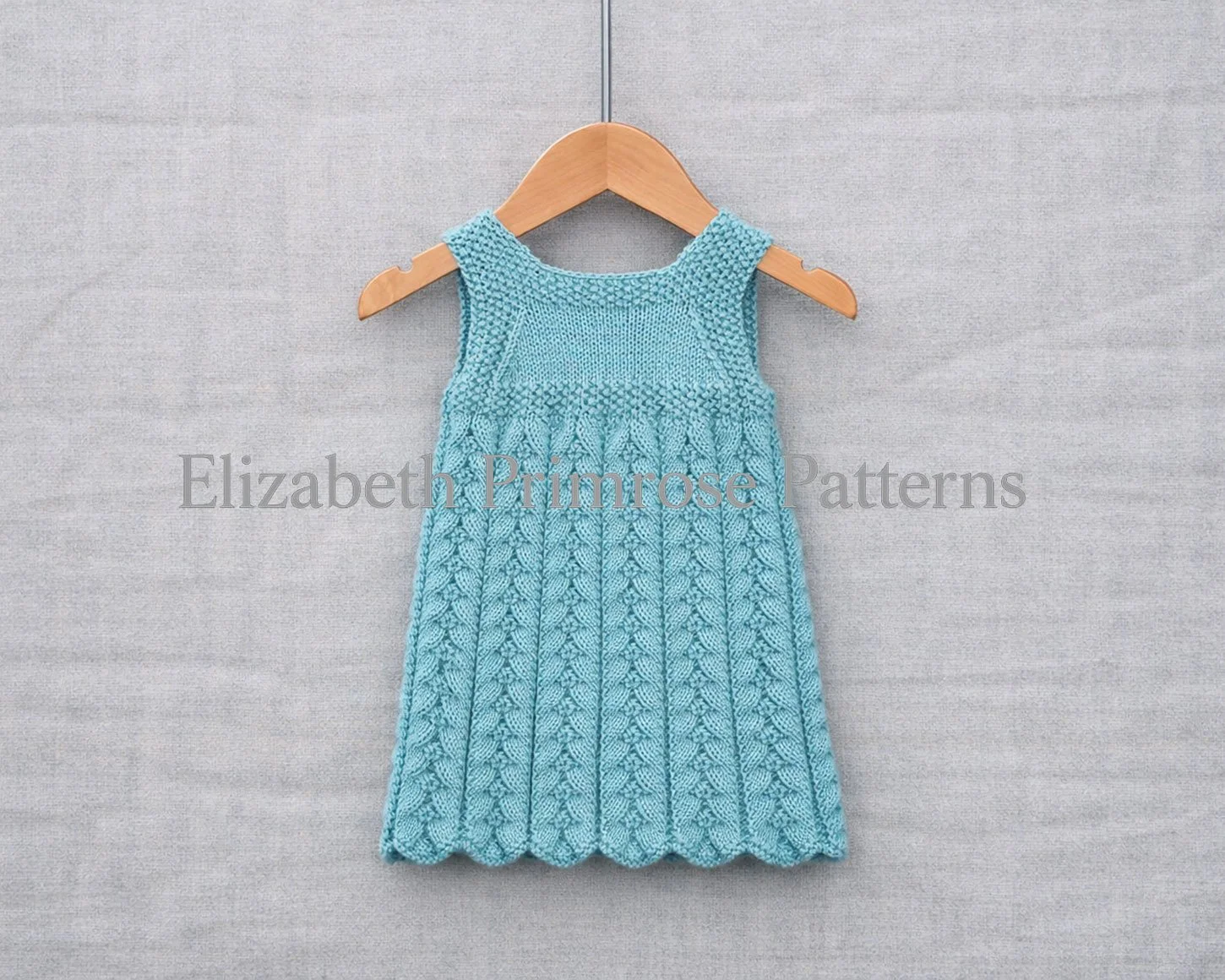 Back view of hand knitted turquoise baby dress on a wooden hanger against a soft fabric backdrop.