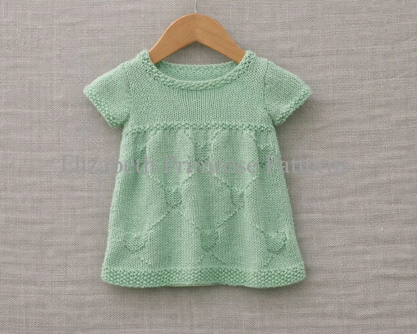 Mint green short-sleeved hand-knitted child dress featuring a textured diamond and heart motif on a wooden hanger.