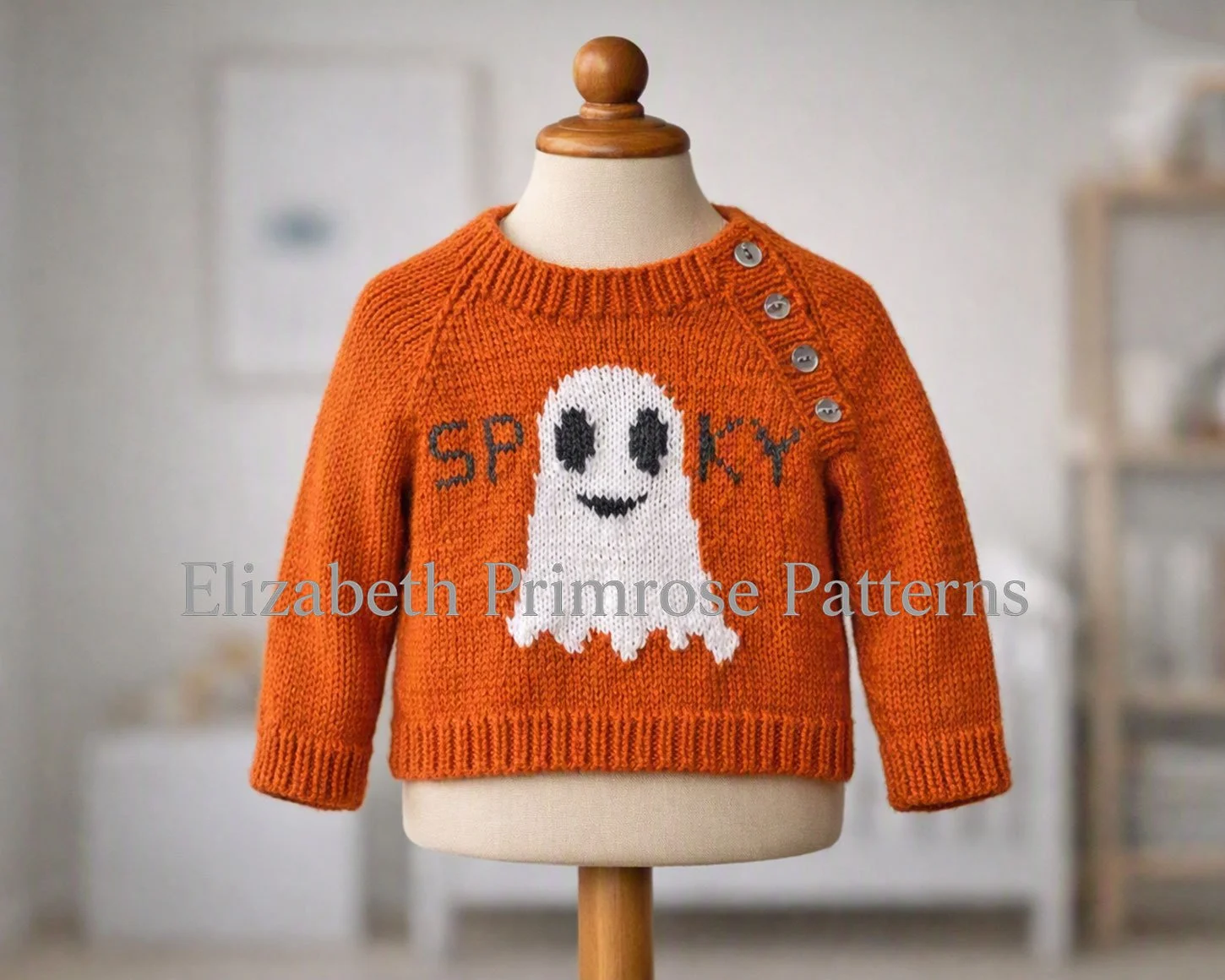 Orange children’s jumper with ghost motif and “spooky” lettering, shown on a mannequin with side buttons in a softly lit neutral room.