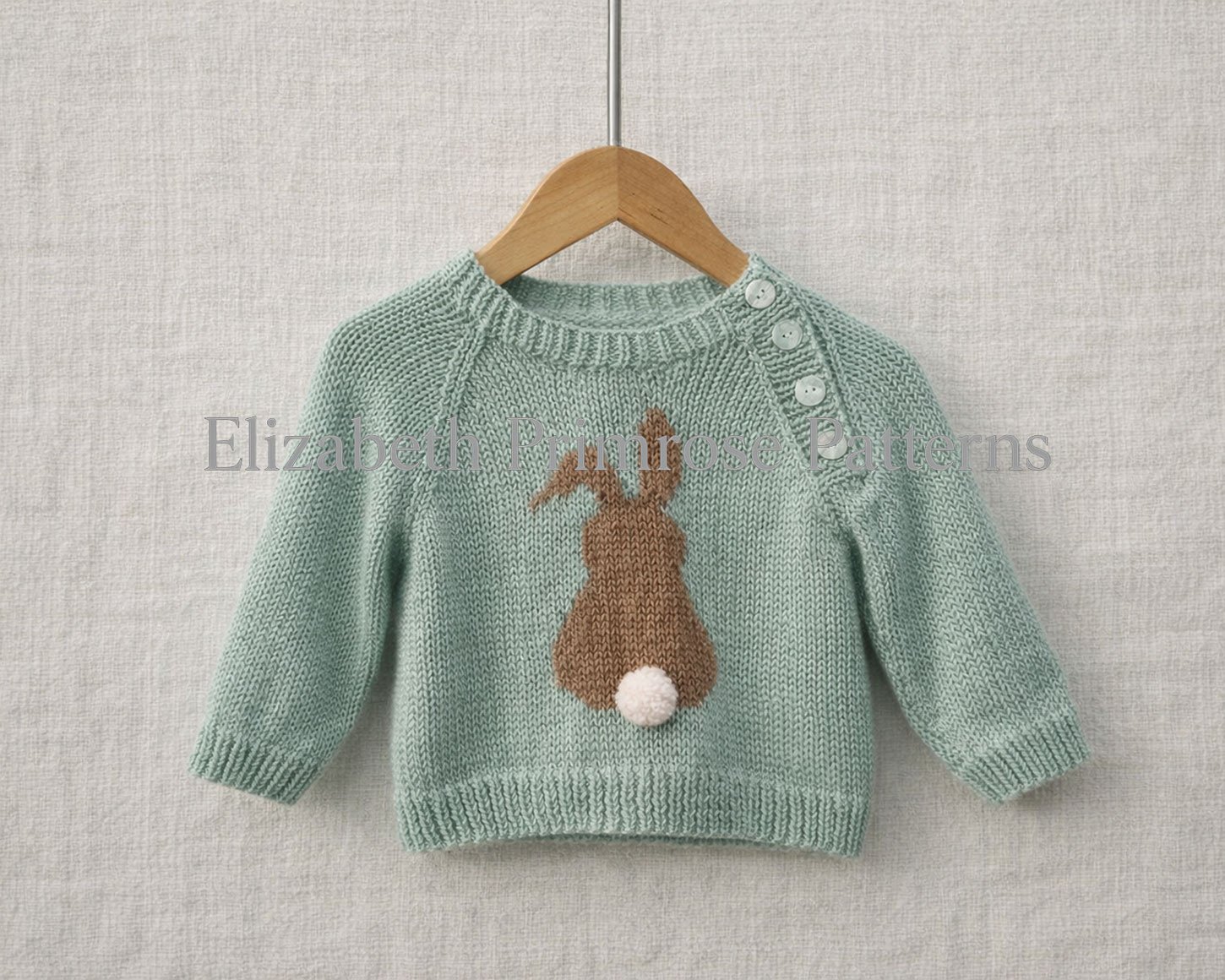 Light green hand‑knitted baby jumper with brown bunny motif and white pom‑pom tail, hanging on wooden hanger against natural fabric background.