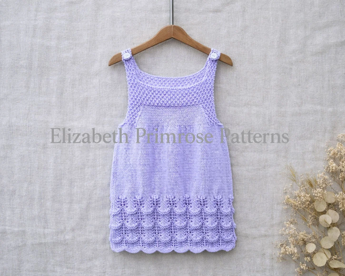 Lilac hand knitted baby dress with textured bodice and scalloped lace hem hanging on a wooden hanger against a soft linen backdrop.