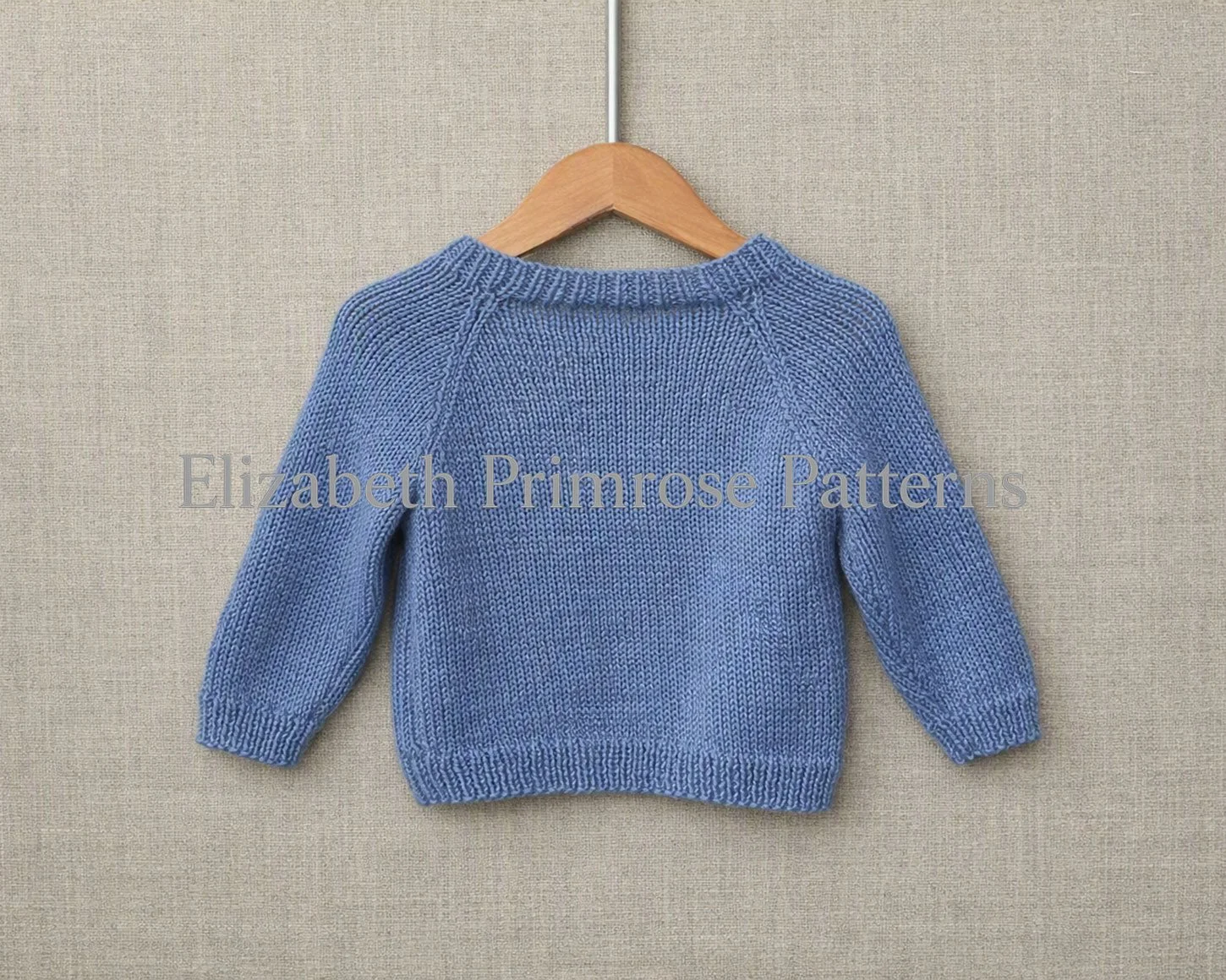 Back view of blue children’s jumper with raglan sleeves hanging on a wooden hanger against a soft beige fabric background.
