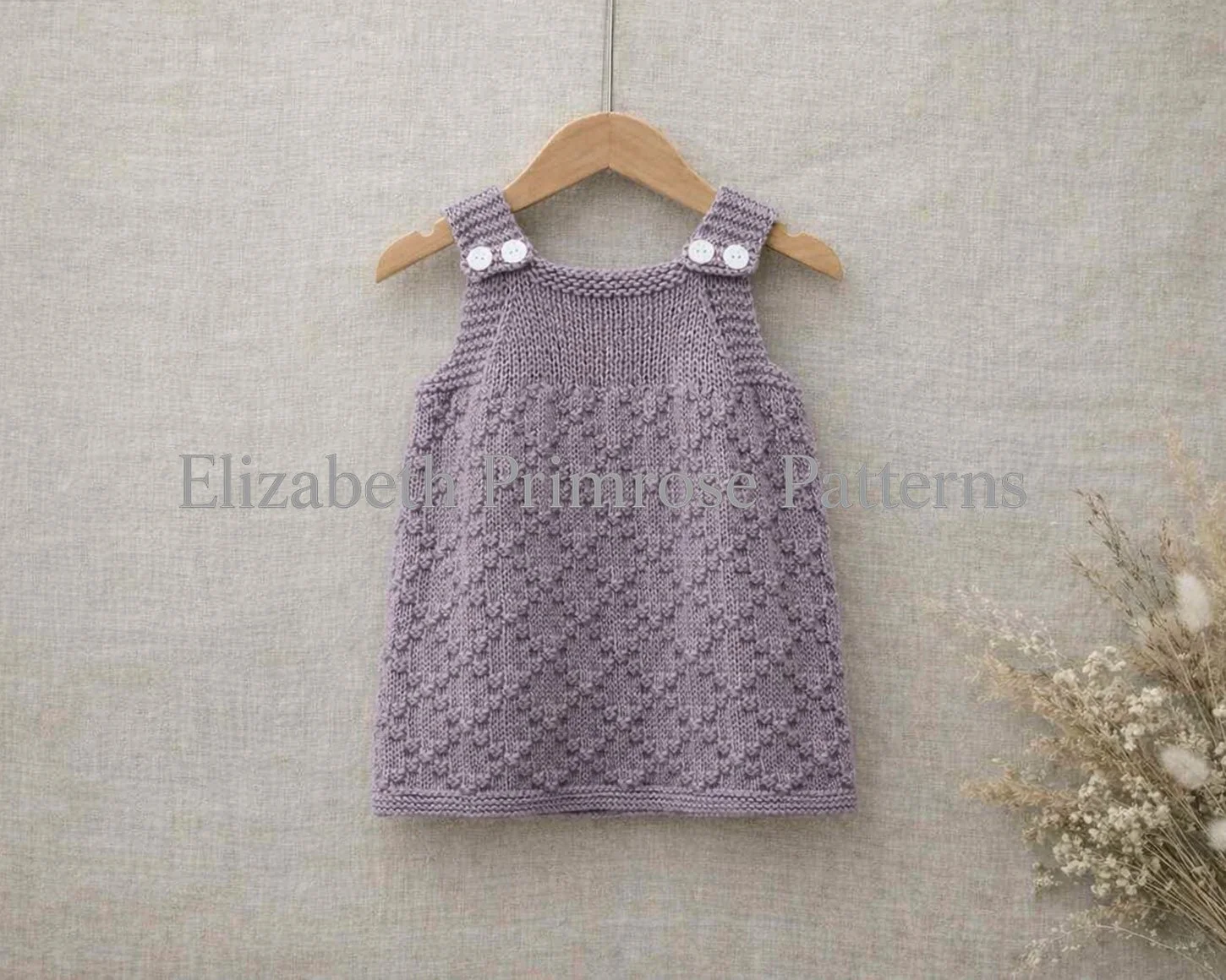 Children’s lavender knitted pinafore dress with diamond texture laid flat on beige linen with soft dried florals beside it.