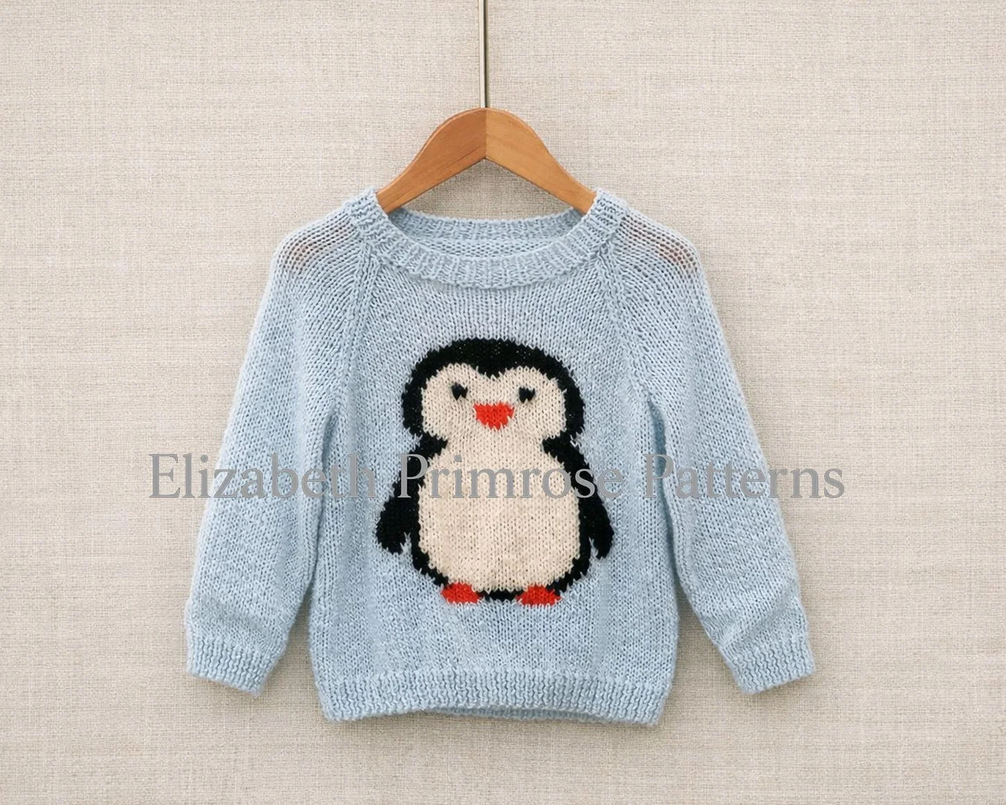 Light blue hand‑knitted jumper with a penguin motif hanging on a wooden hanger against a beige linen background.
