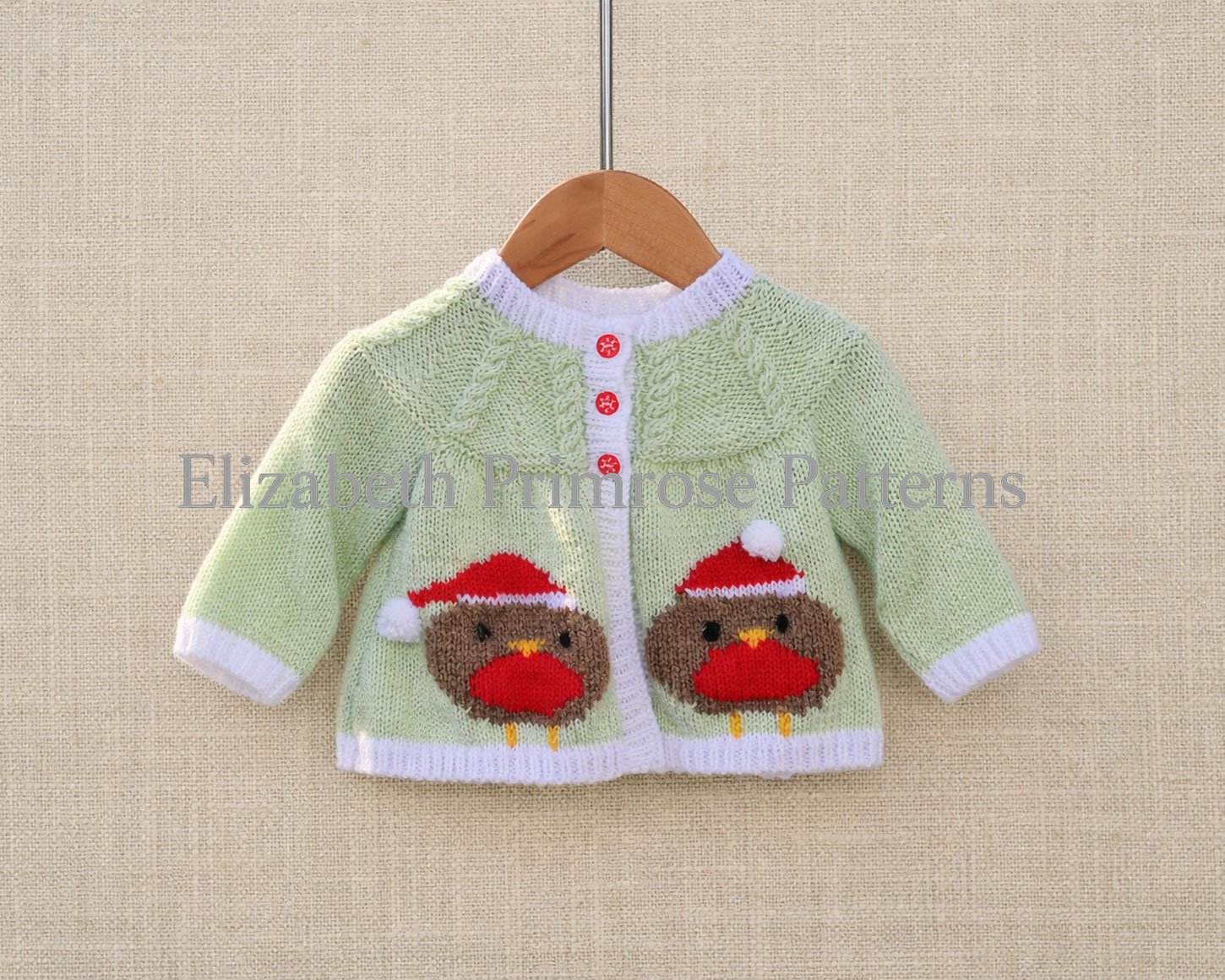 Light green baby cardigan with two Christmas robin motifs hanging on a wooden hanger against a soft beige fabric background.