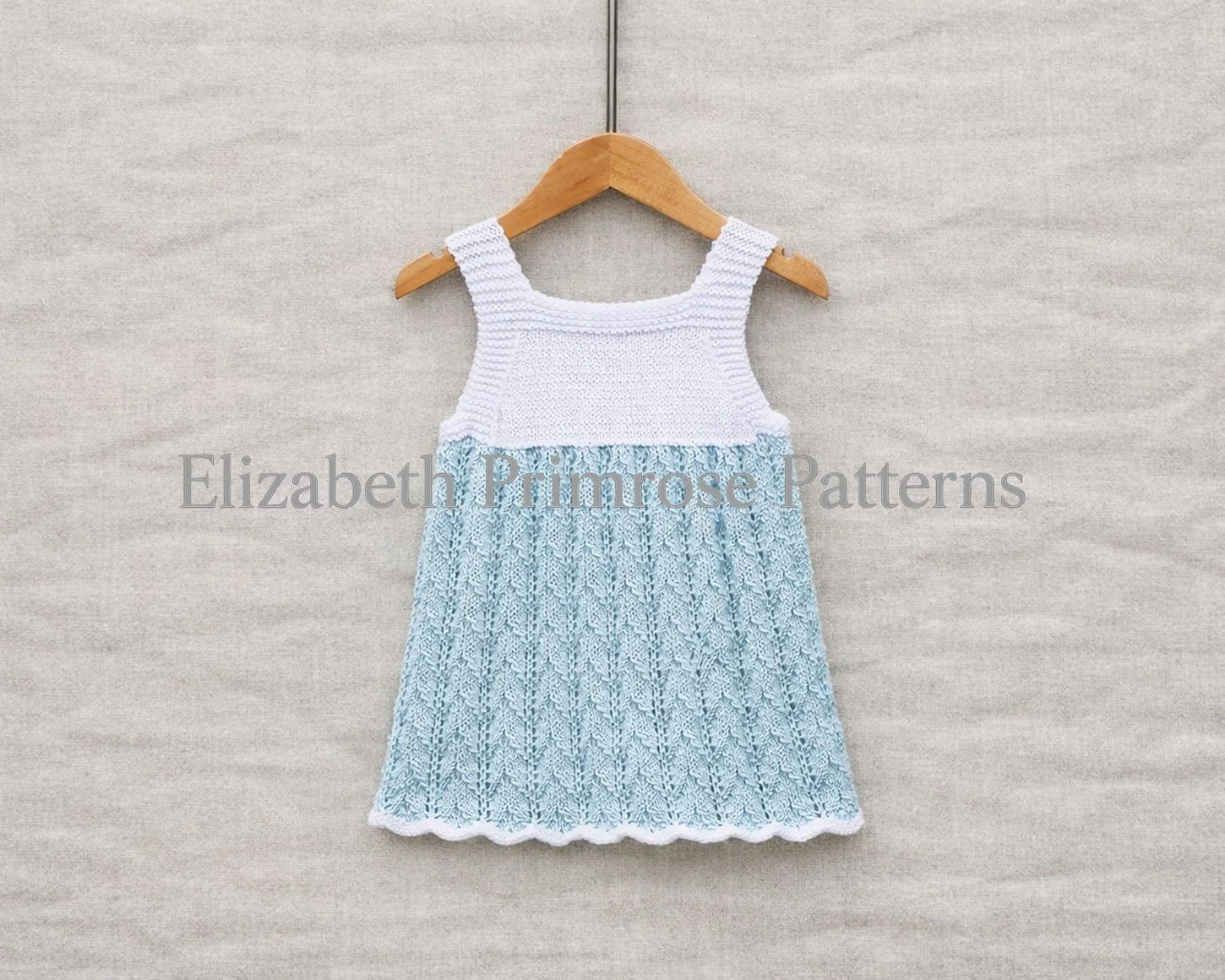 Back view of hand knitted baby sundress displayed on hanger with scalloped hem on a neutral fabric background.