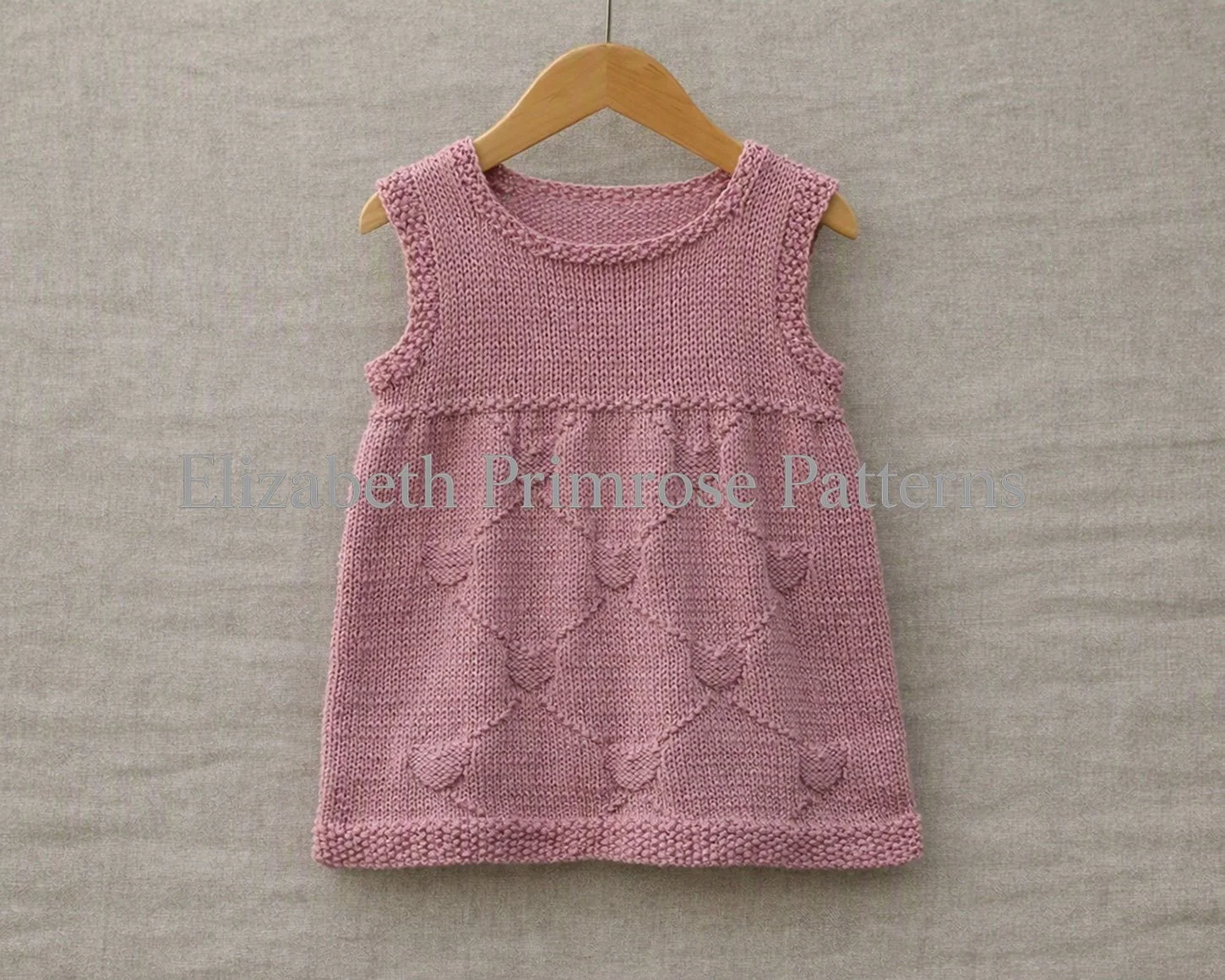 Dusty pink sleeveless hand-knitted child dress featuring a textured diamond and heart motif on a wooden hanger.