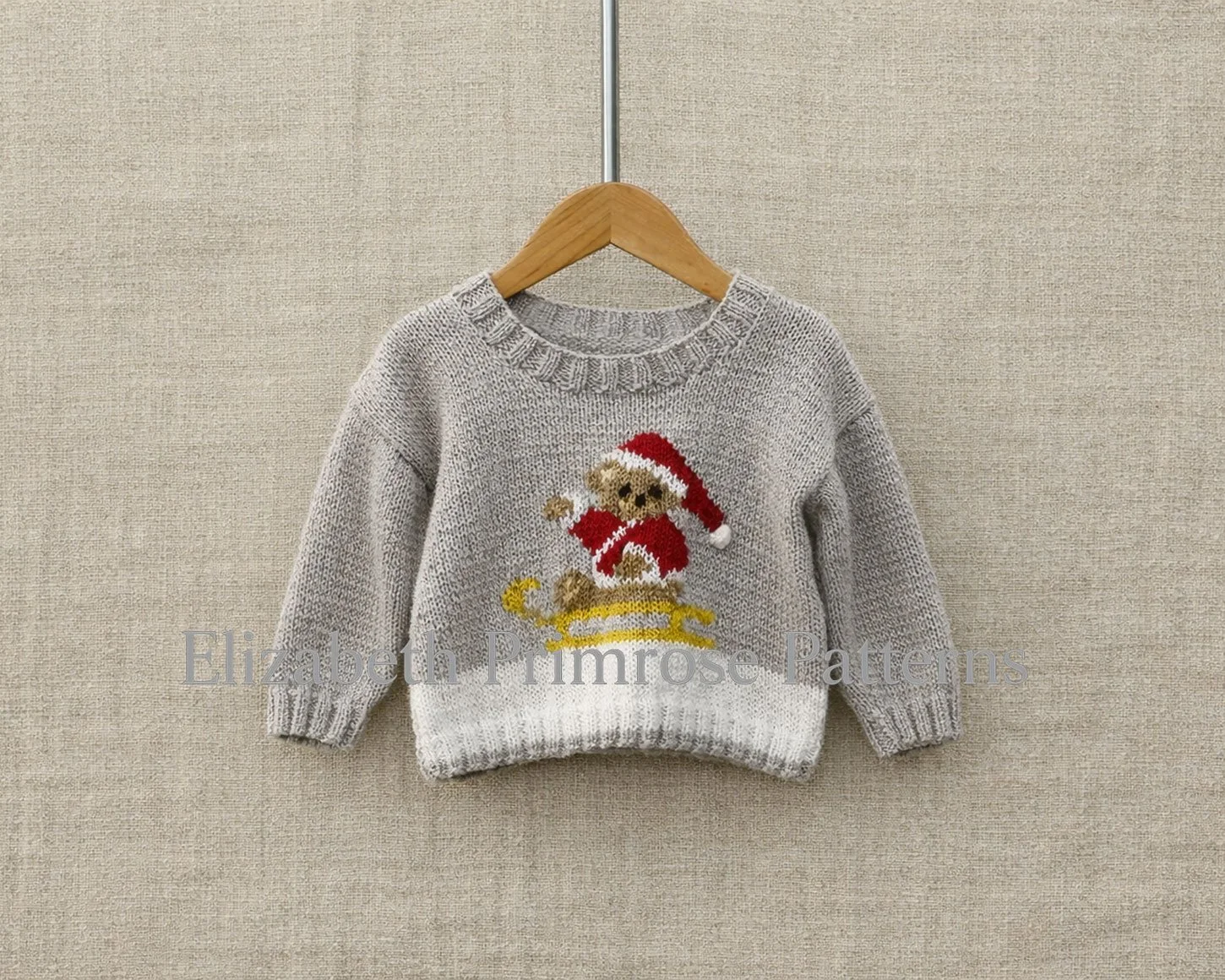 Grey children’s jumper with Santa teddy bear on a yellow sleigh, hanging on a wooden hanger against a beige linen background.