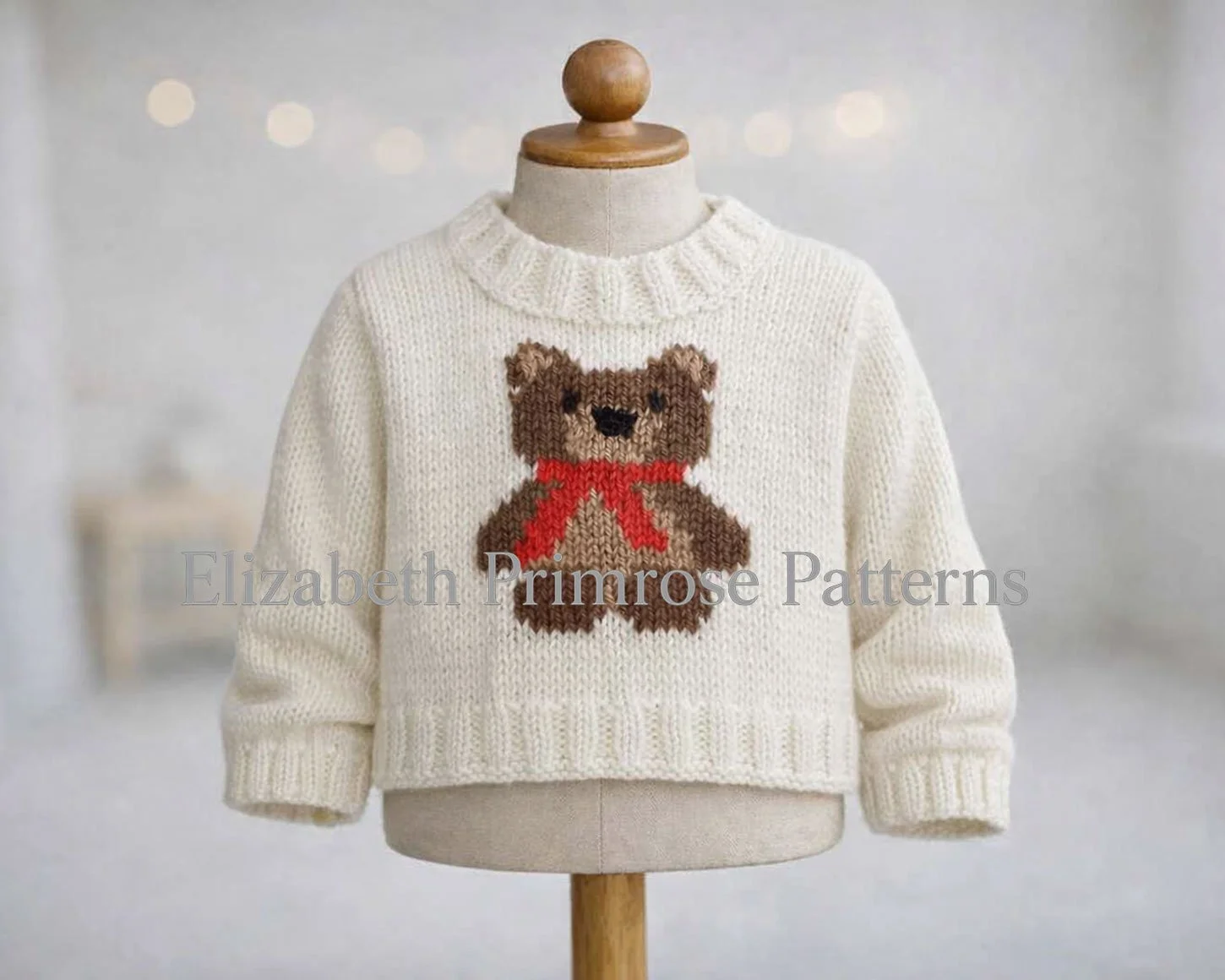 Cream knitted children’s jumper on a mannequin, featuring a brown teddy bear with a red scarf. Soft, bright room background.