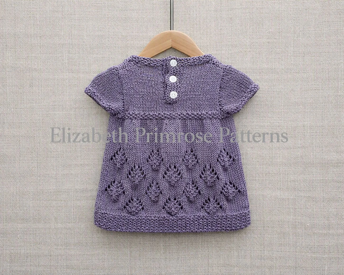 Lavender knitted children’s dress with pinecone pattern skirt, sand stitch hem and buttons hanging from a wooden hanger.