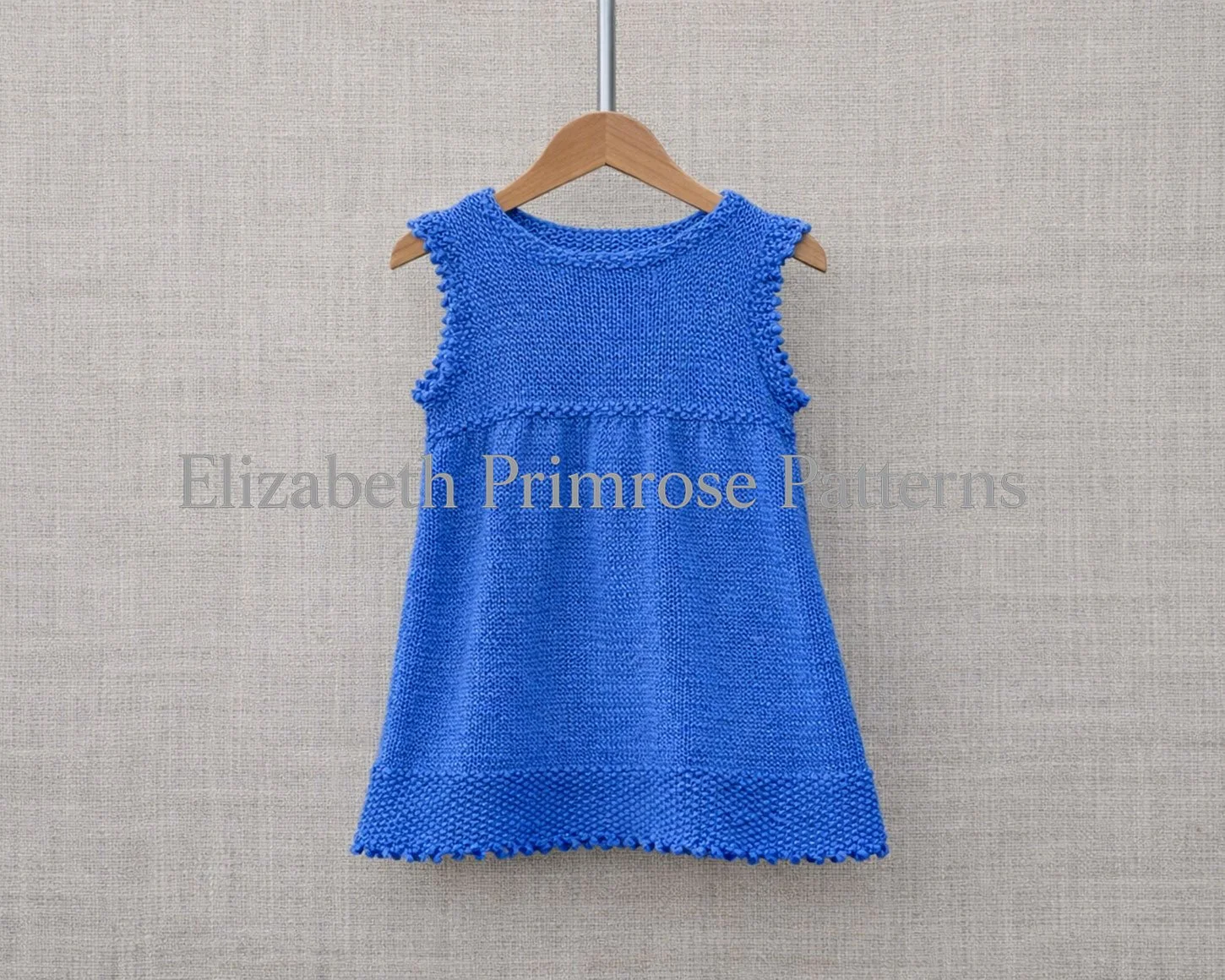 Blue hand‑knitted children’s dress hanging on a wooden hanger against a textured linen background.