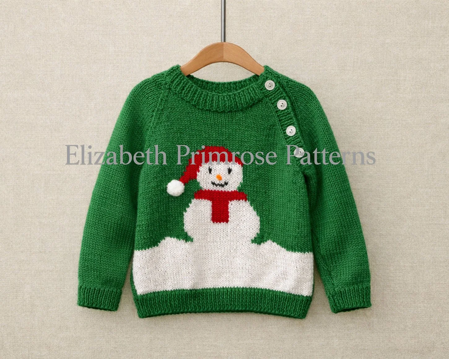 Green children’s snowman jumper hanging on a wooden hanger against a soft beige background, showing buttoned shoulder and festive snowman motif.