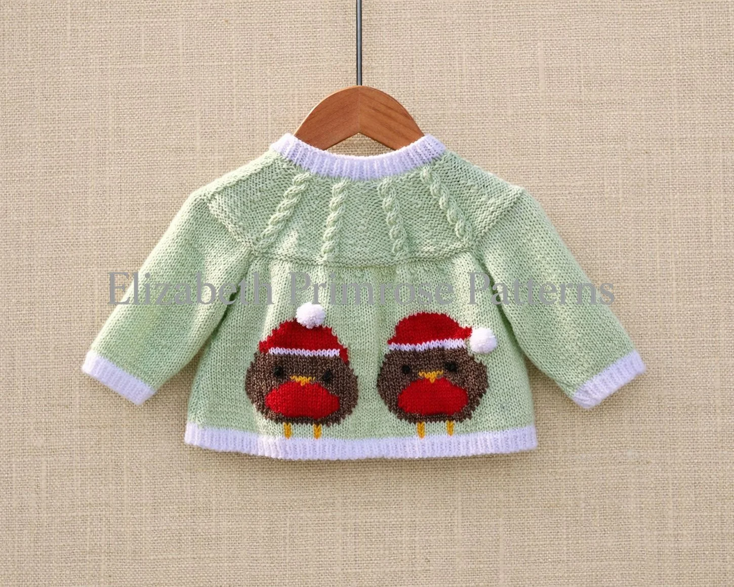 Back of knitted baby jacket with festive robin motifs, cabled yoke and white edging, hanging on a wooden hanger over a neutral fabric backdrop.
