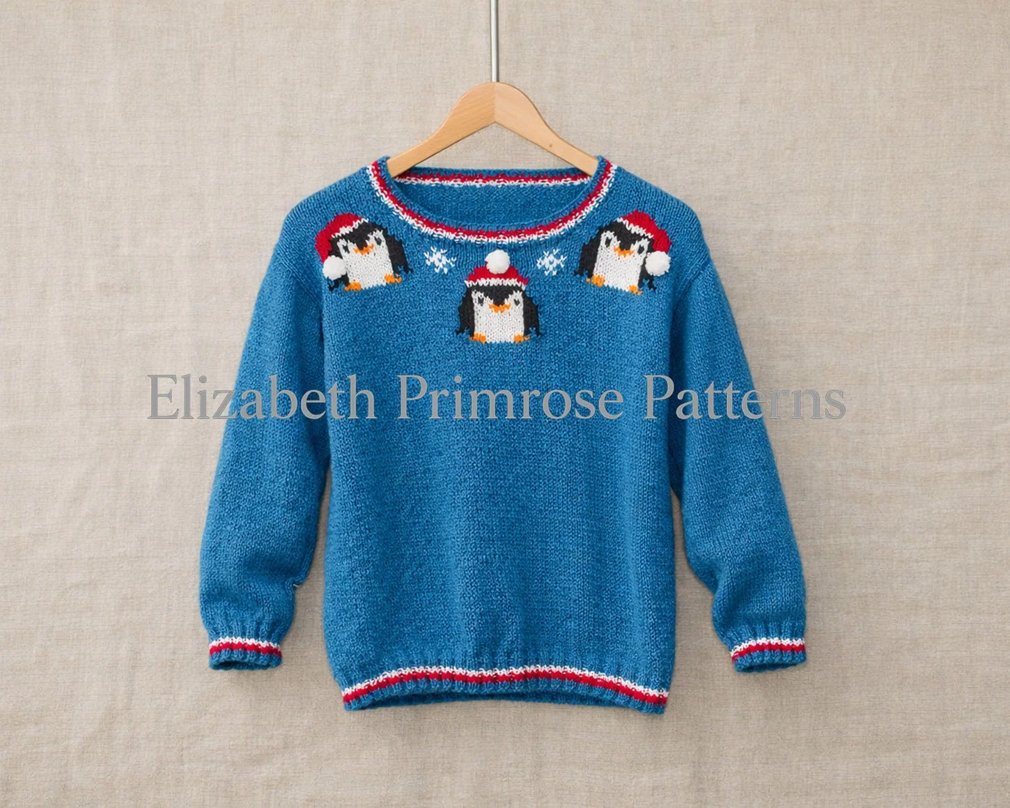 Blue children’s jumper with three Christmas penguins in Santa hats, hanging on a wooden hanger against a beige linen background.