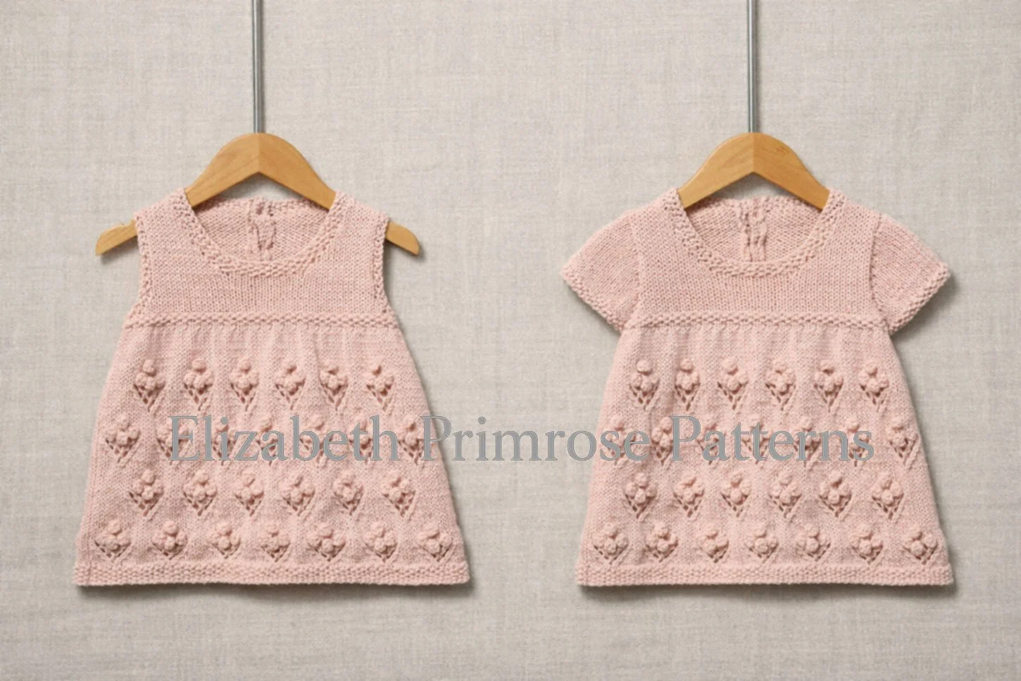 Light pink knitted baby dresses with bobble flower skirts hanging on wooden hangers, showing sleeveless and short sleeved styles against a soft fabric backdrop.