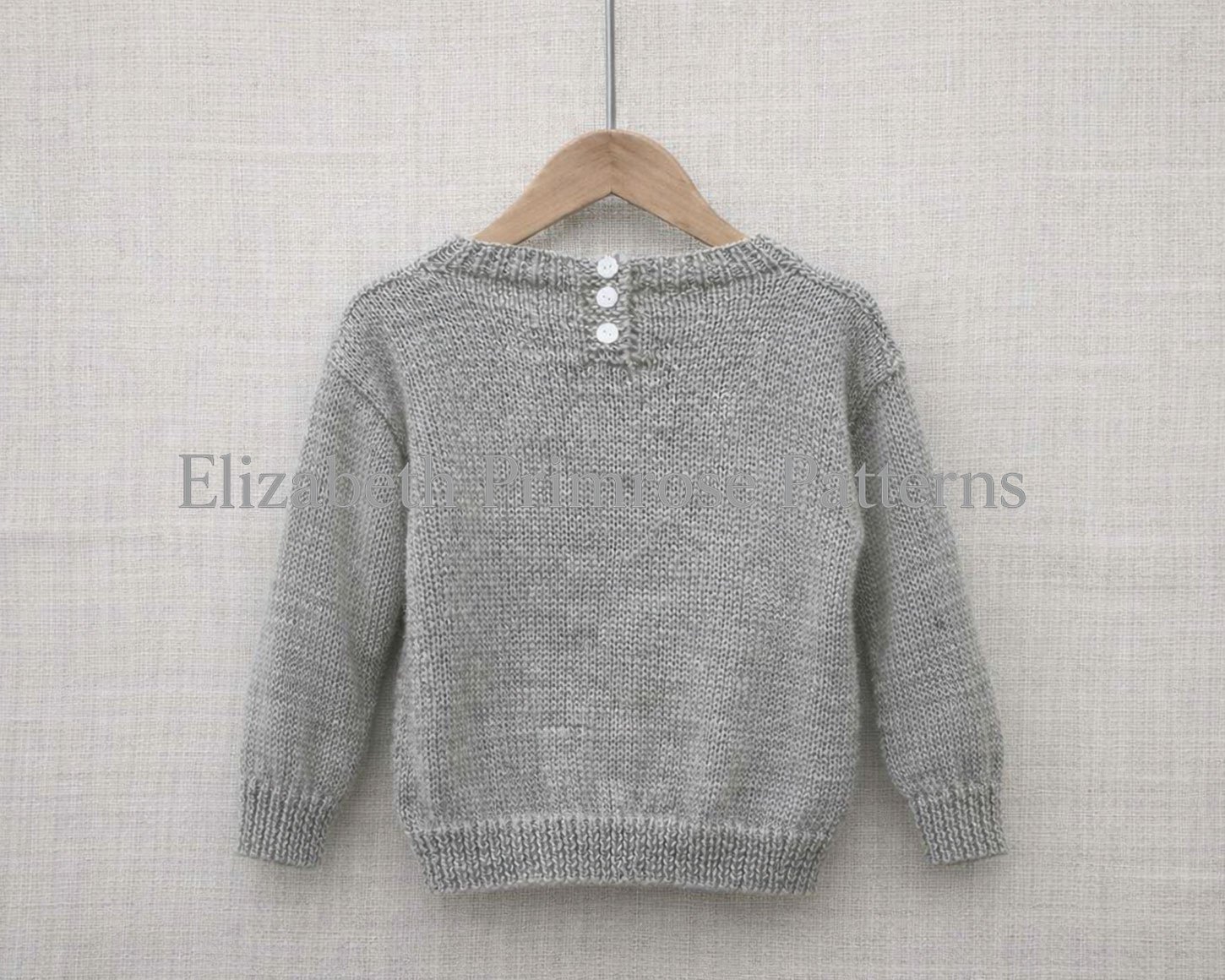 Back view of hand‑knitted grey‑green children’s jumper with three button fastening, hanging on a wooden hanger against a light linen backdrop.