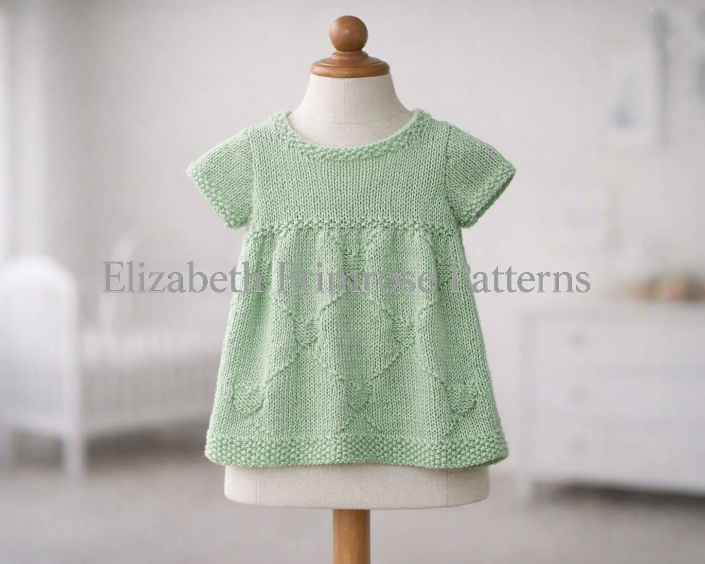 Hand-knitted mint green toddler dress on a neutral wooden stand mannequin, showcasing the intricate lace motif against a softly blurred nursery bedroom background.