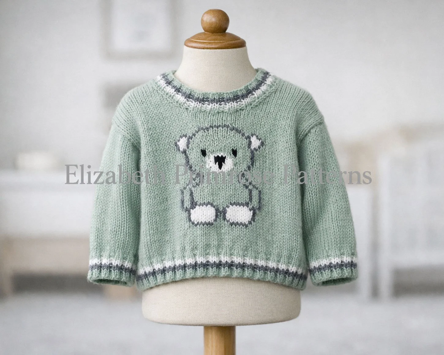 Sage green knitted baby sweater featuring bear design, shown on wooden-topped mannequin with natural sleeve drape and blurred minimalist room backdrop.