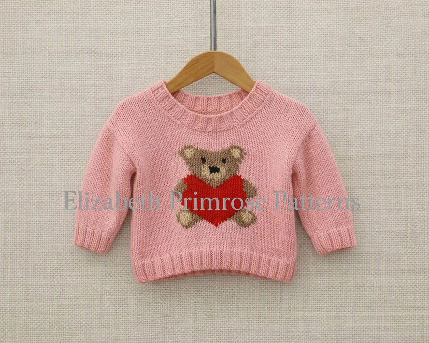 Flat lay view of a pink knitted kids' sweater on a wooden hanger, featuring a textured teddy bear and red heart design.