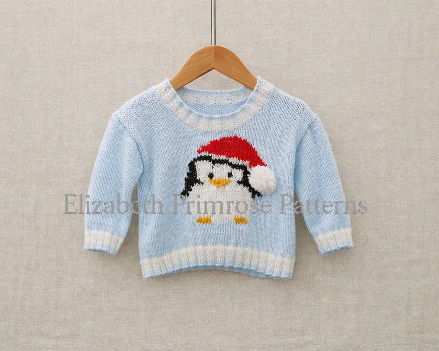 Light blue children’s jumper with penguin in Santa hat, displayed on a wooden hanger against a soft beige fabric background.