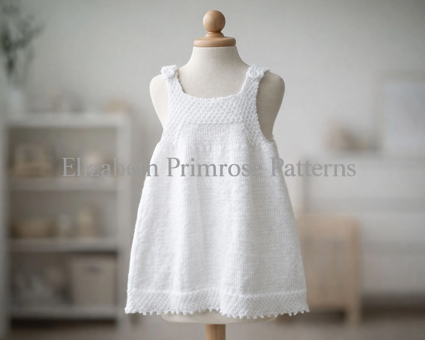 White hand knitted children’s pinafore on a fabric mannequin with buttoned straps, textured bodice and smooth skirt, shown in a softly blurred Scandinavian nursery.